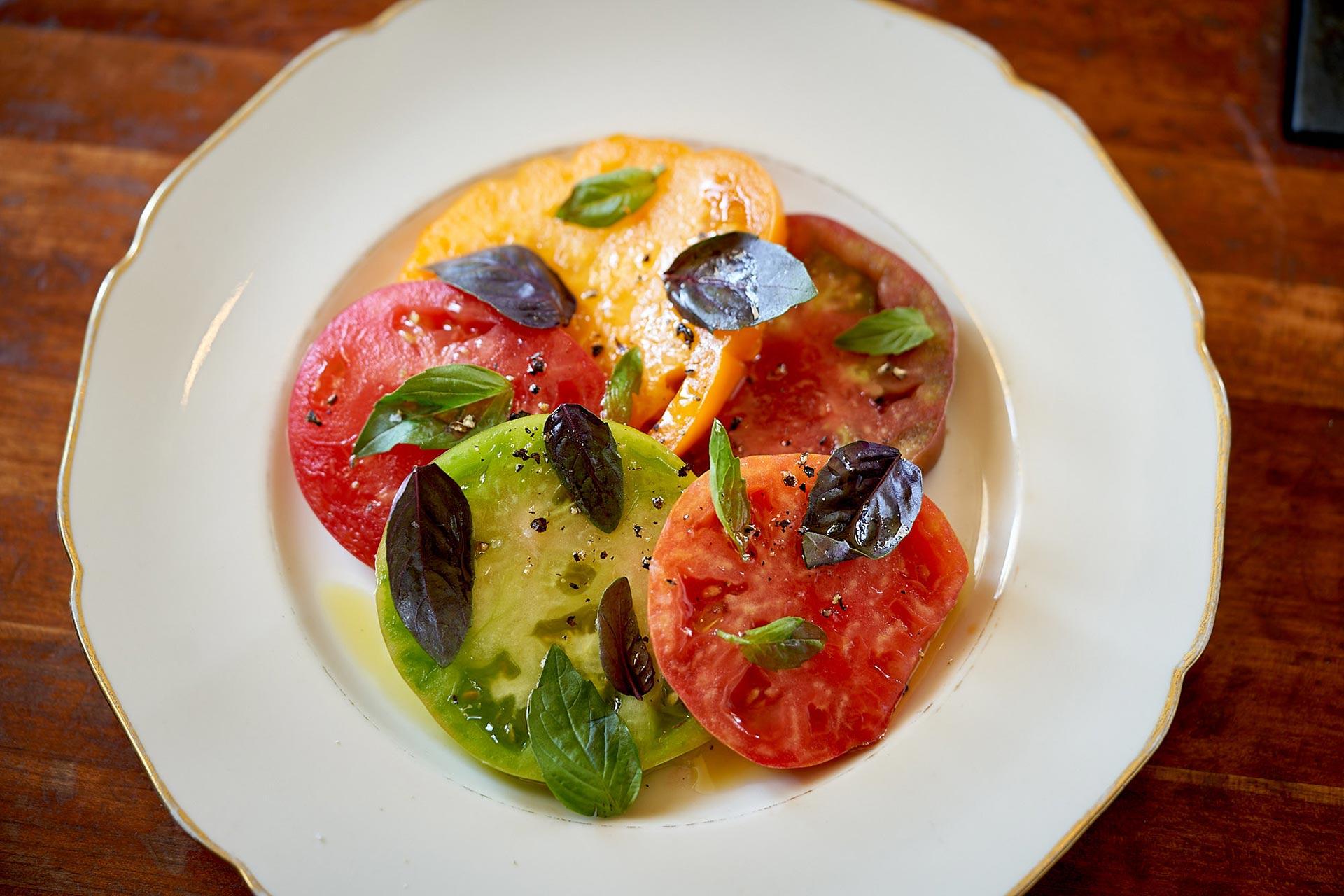 A plate of colorful heirloom tomato slices topped with fresh basil leaves and cracked black pepper, served on a white dish with a gold rim, on a wooden table.
