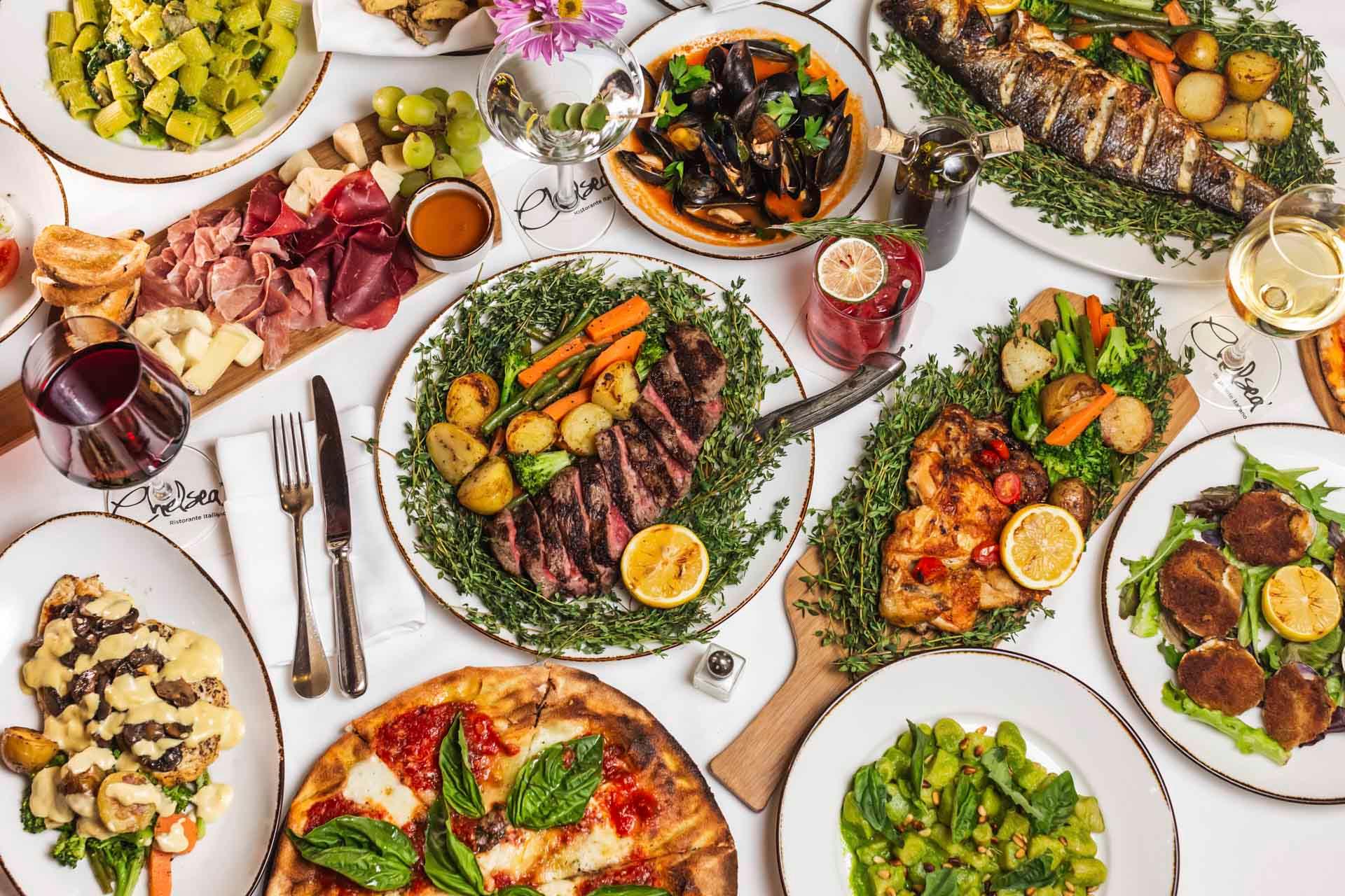 A lavish spread of Italian cuisine featuring dishes like pizza, steak with vegetables, grilled fish, and assorted cheeses. Accompanied by wine and garnished with fresh herbs, the table displays a vibrant and appetizing array of meals.