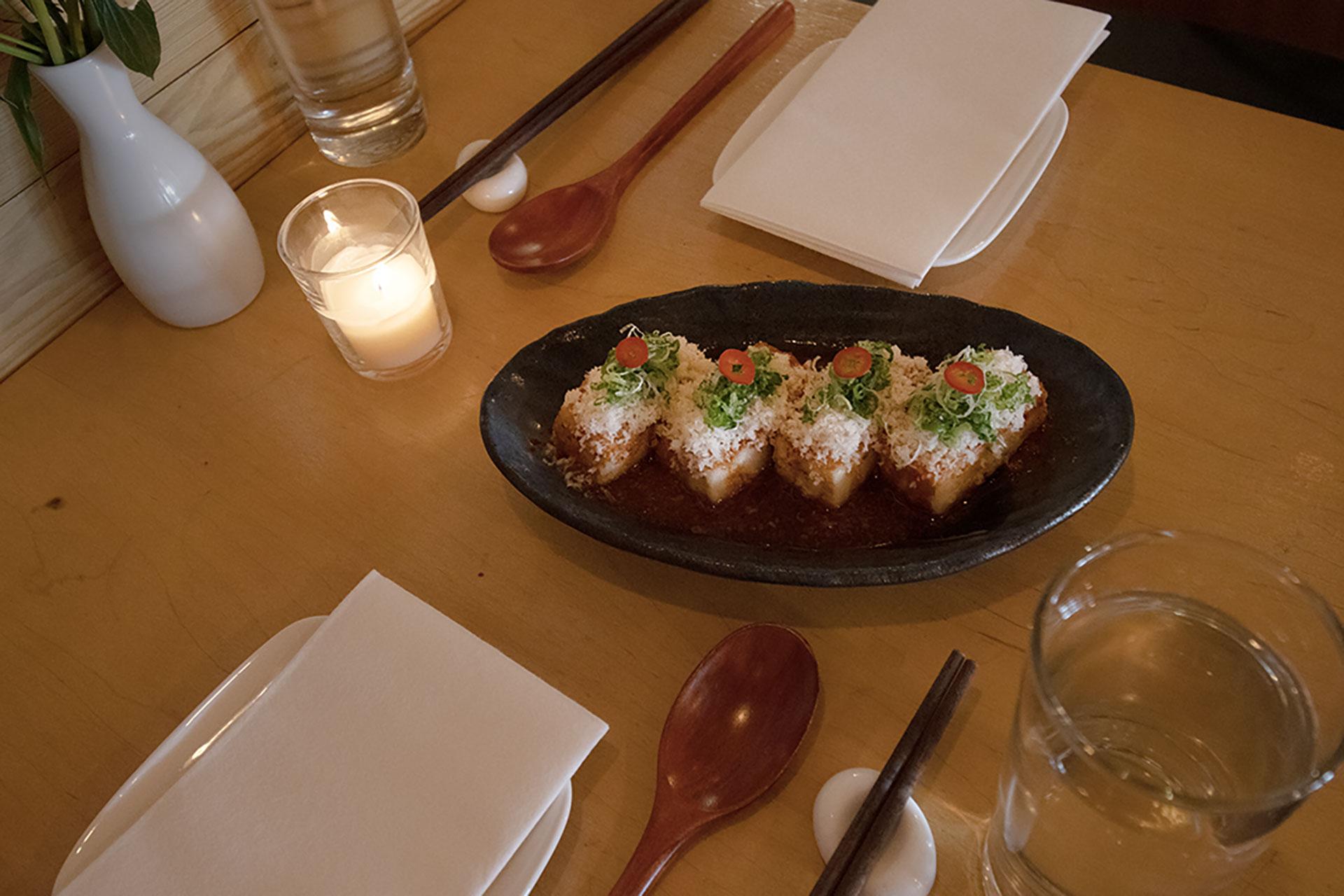 A black oval plate holds four pieces of crispy rice topped with green onions, sauce, and garnishes on a wooden table set with glassware, utensils, napkins, and a lit candle.