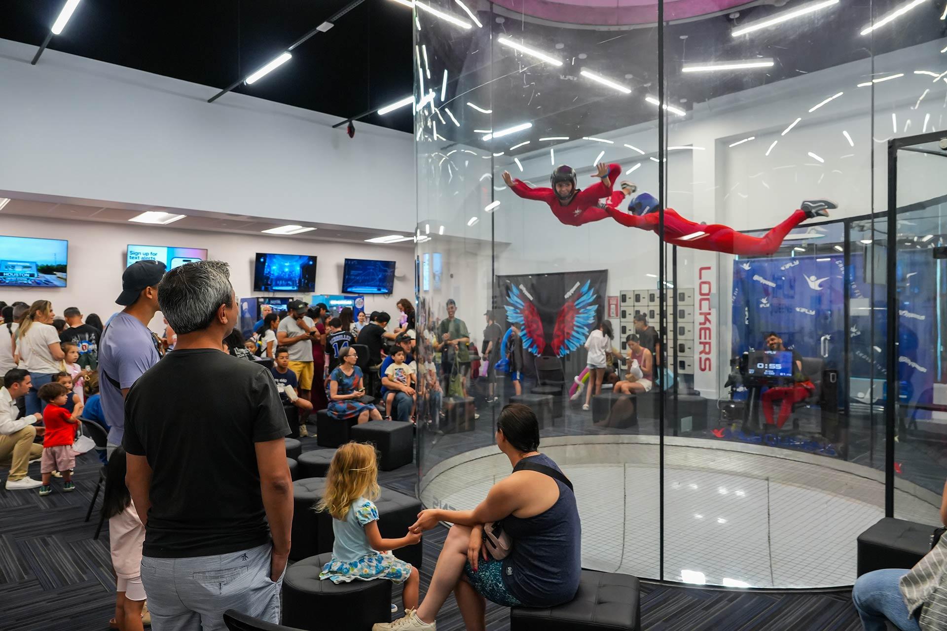 A crowd watches as two people in red suits fly inside an indoor skydiving wind tunnel. Some children and adults sit and stand near the glass, observing the demonstration in a modern, well-lit facility.