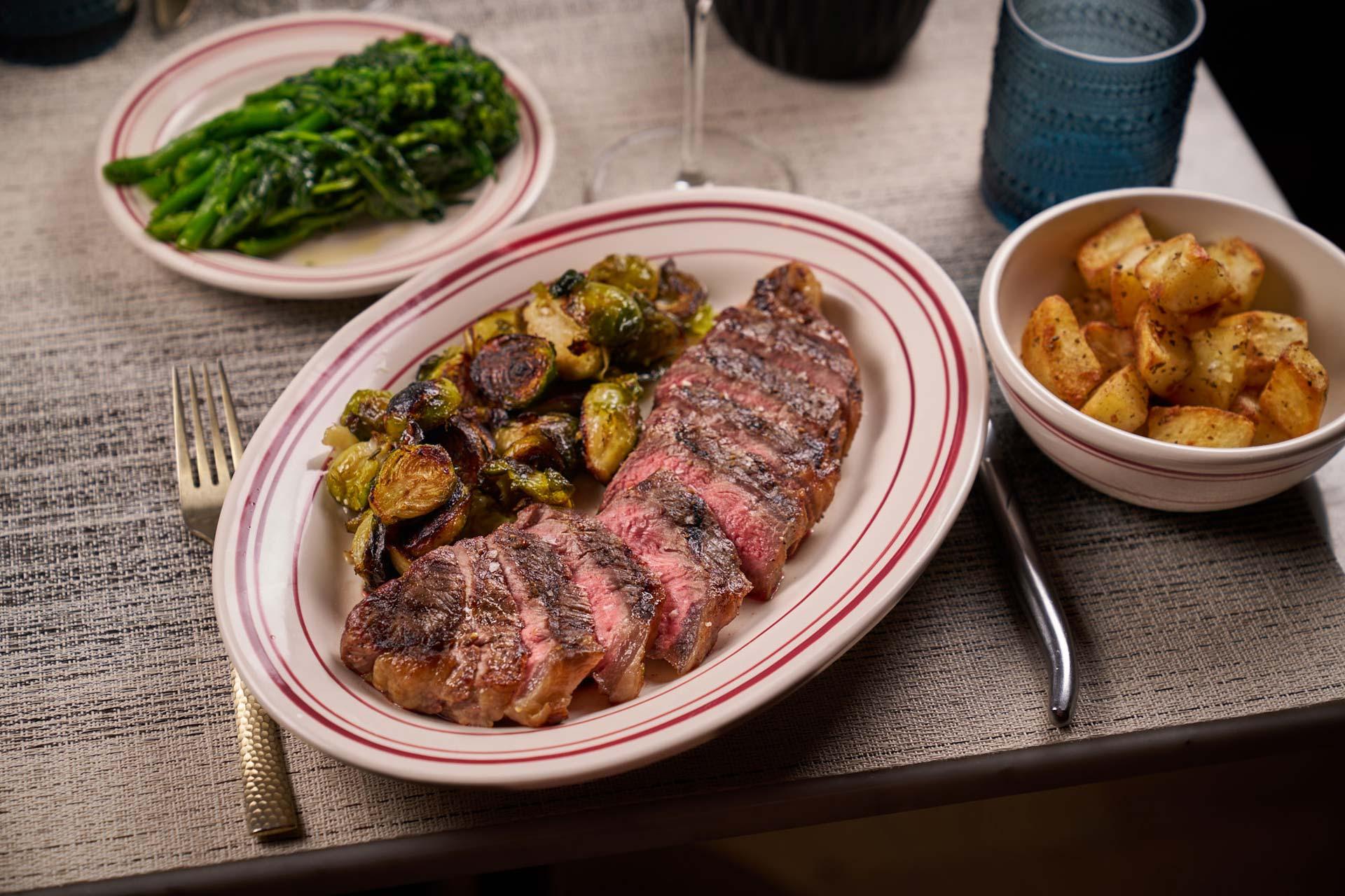 A plate with sliced steak and roasted Brussels sprouts, served with a side dish of seasoned potatoes and a plate of sautéed broccolini. Blue glasses and a fork are on the table.
