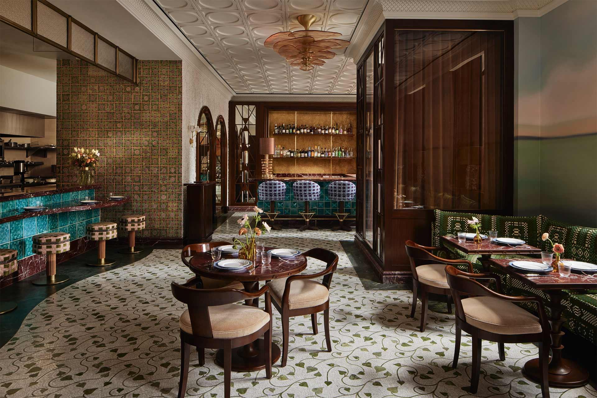 Elegant restaurant interior with patterned floor, wooden tables, cushioned chairs, green banquette seating, a teal-tiled bar, bar stools, and decorative lighting. Shelves with bottles are visible behind the bar area.