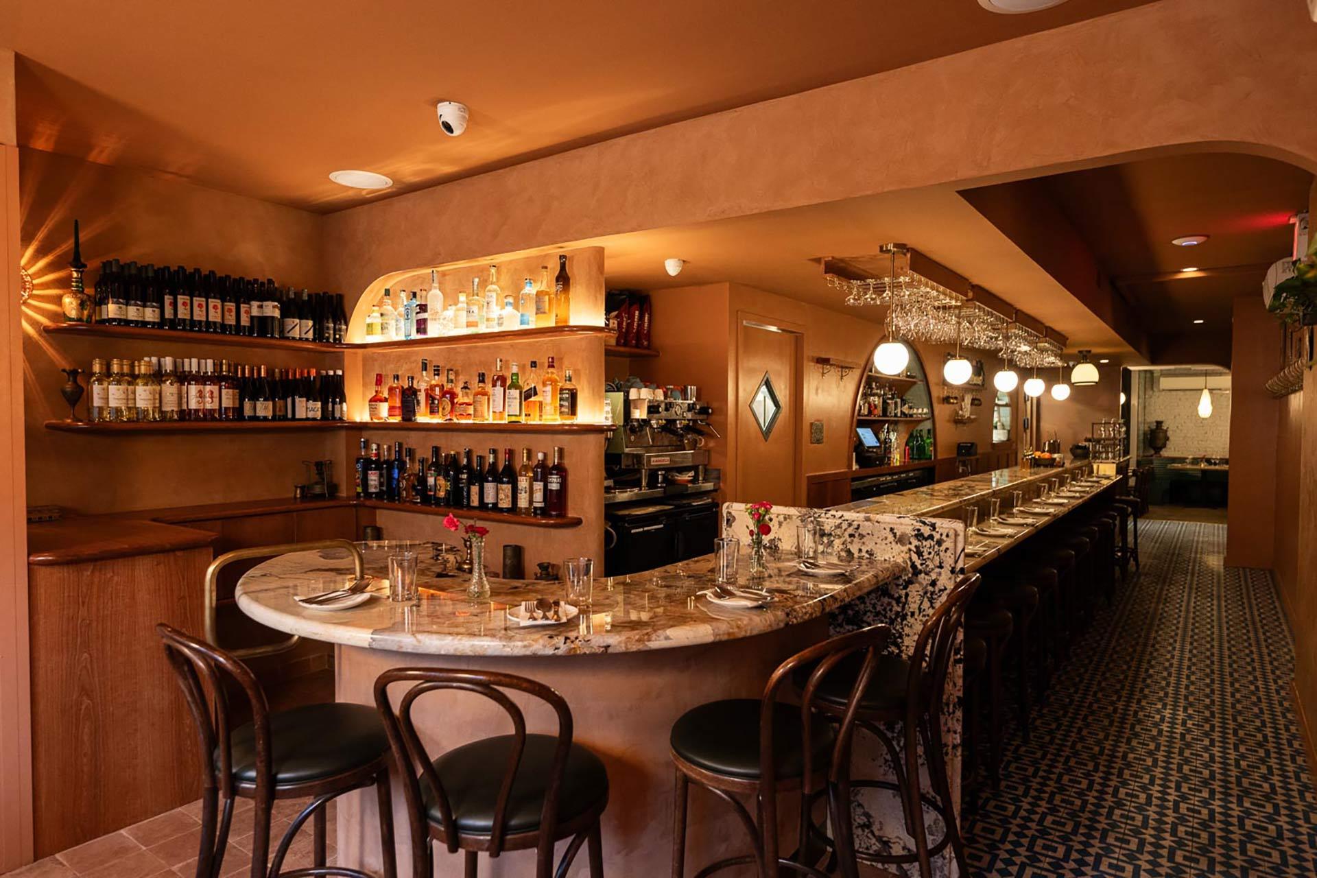 Cozy restaurant bar with curved marble counter, wooden chairs, shelves of bottles, and hanging glasses. Warm lighting, patterned tile floor, and tables set for dining create an inviting atmosphere.