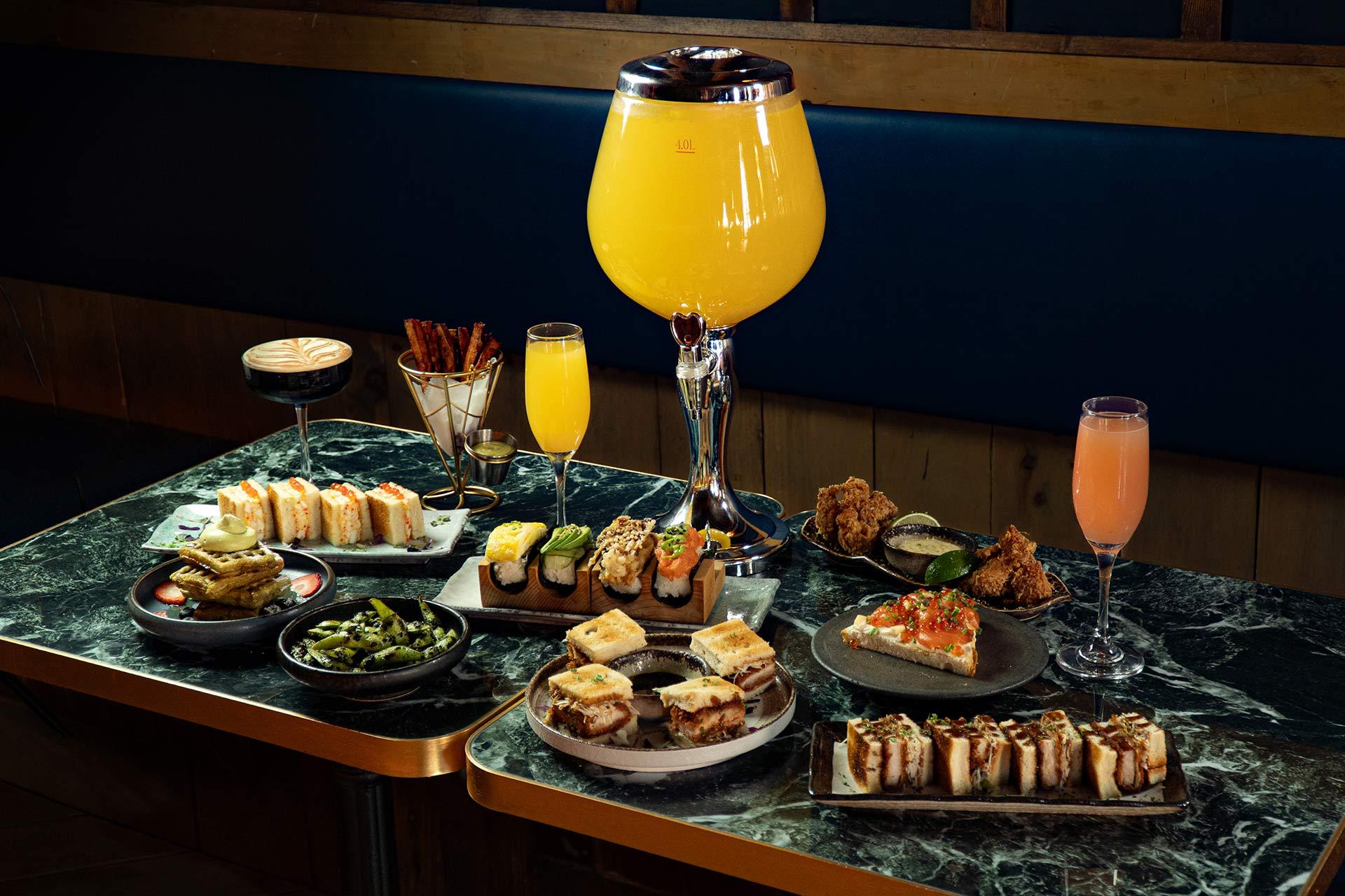 A marble table filled with assorted Japanese small plates, sandwiches, fried chicken, edamame, cocktails, and a large glass dispenser of orange drink in the center. The setting is modern and stylish.