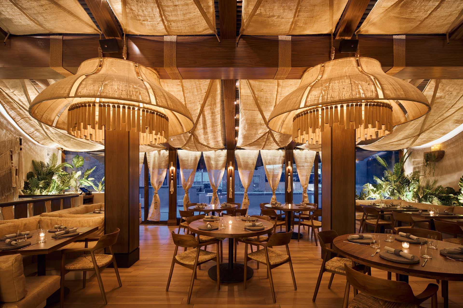 Elegant restaurant interior with large, sculptural pendant lights, wooden tables and chairs, beige fabric ceiling, soft lighting, and lush green plants along the windows, creating a warm and inviting dining atmosphere.