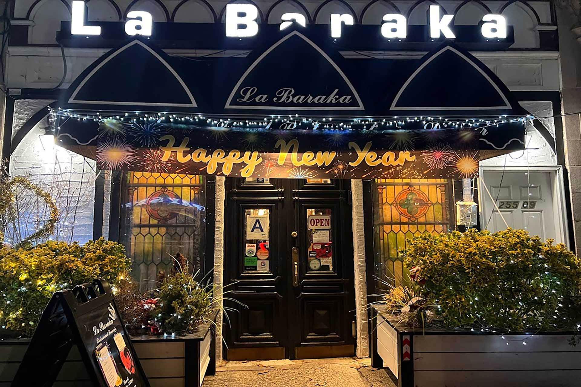 Front of La Baraka restaurant decorated with "Happy New Year" banner and lights. Two stained glass windows flank the entrance. An "A" rating and "Open" sign are on the door. Plants are on either side of the entrance. Nighttime setting.