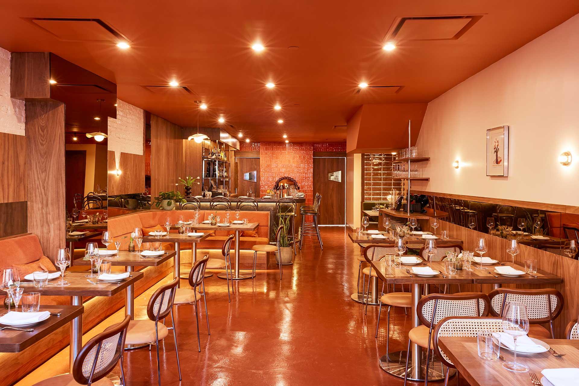 A warmly lit restaurant interior with wooden tables, modern chairs, place settings, and cushioned booths. The décor features earthy tones, exposed brick, and plants, creating a cozy and inviting atmosphere.
