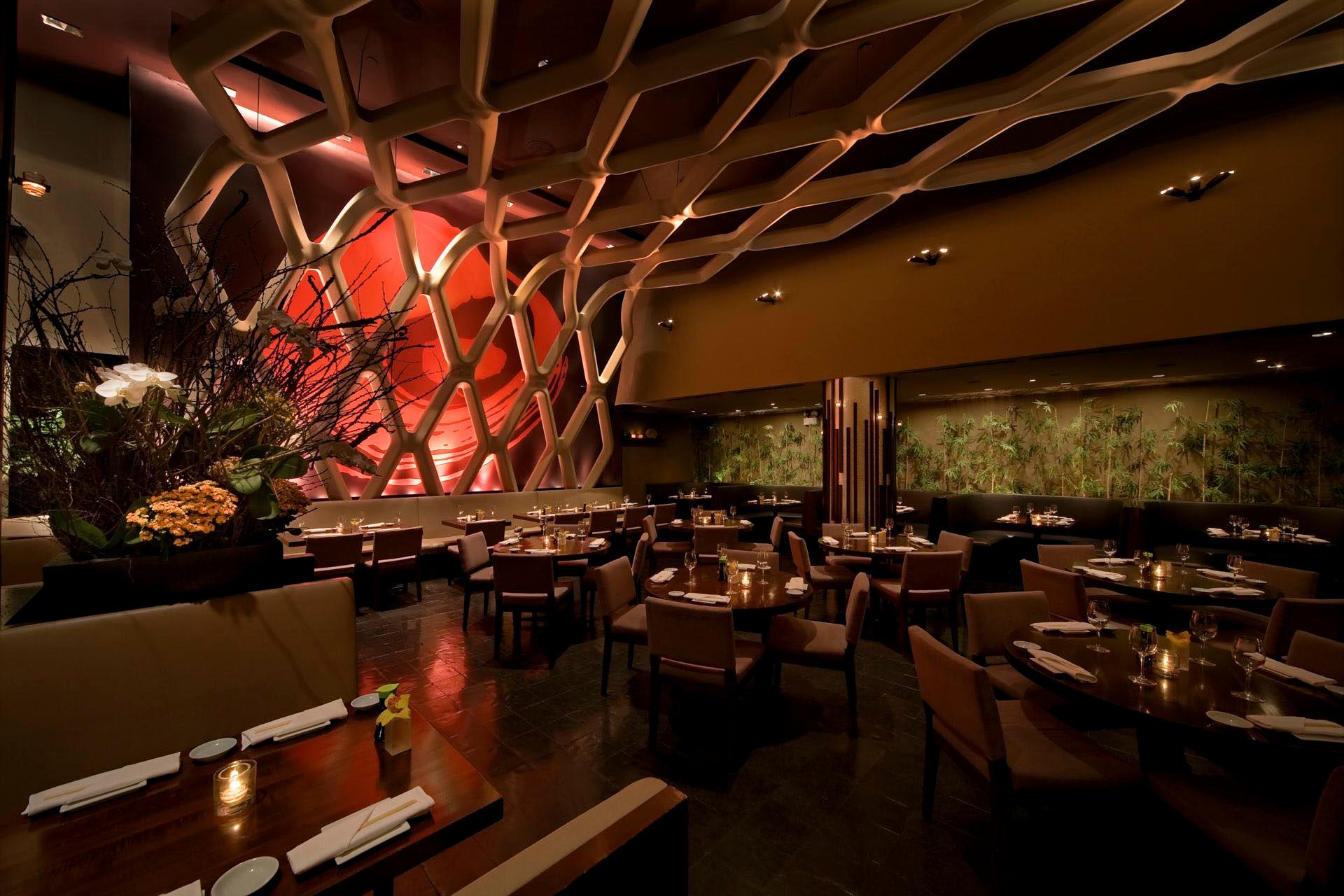 Dimly lit, elegant restaurant interior featuring contemporary decor with a geometric wall design, round wooden tables set with white napkins, and cushioned chairs. Floral arrangements enhance the ambiance.