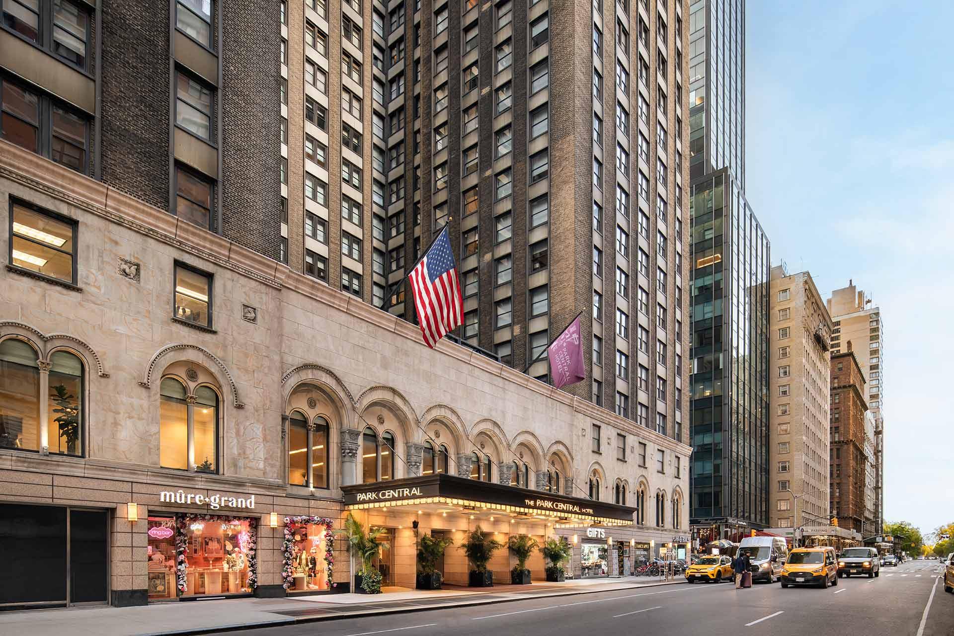 Street view of a tall hotel building with American flags, shops including "mure+grand," and yellow taxis parked along the curb in a busy city, likely New York.