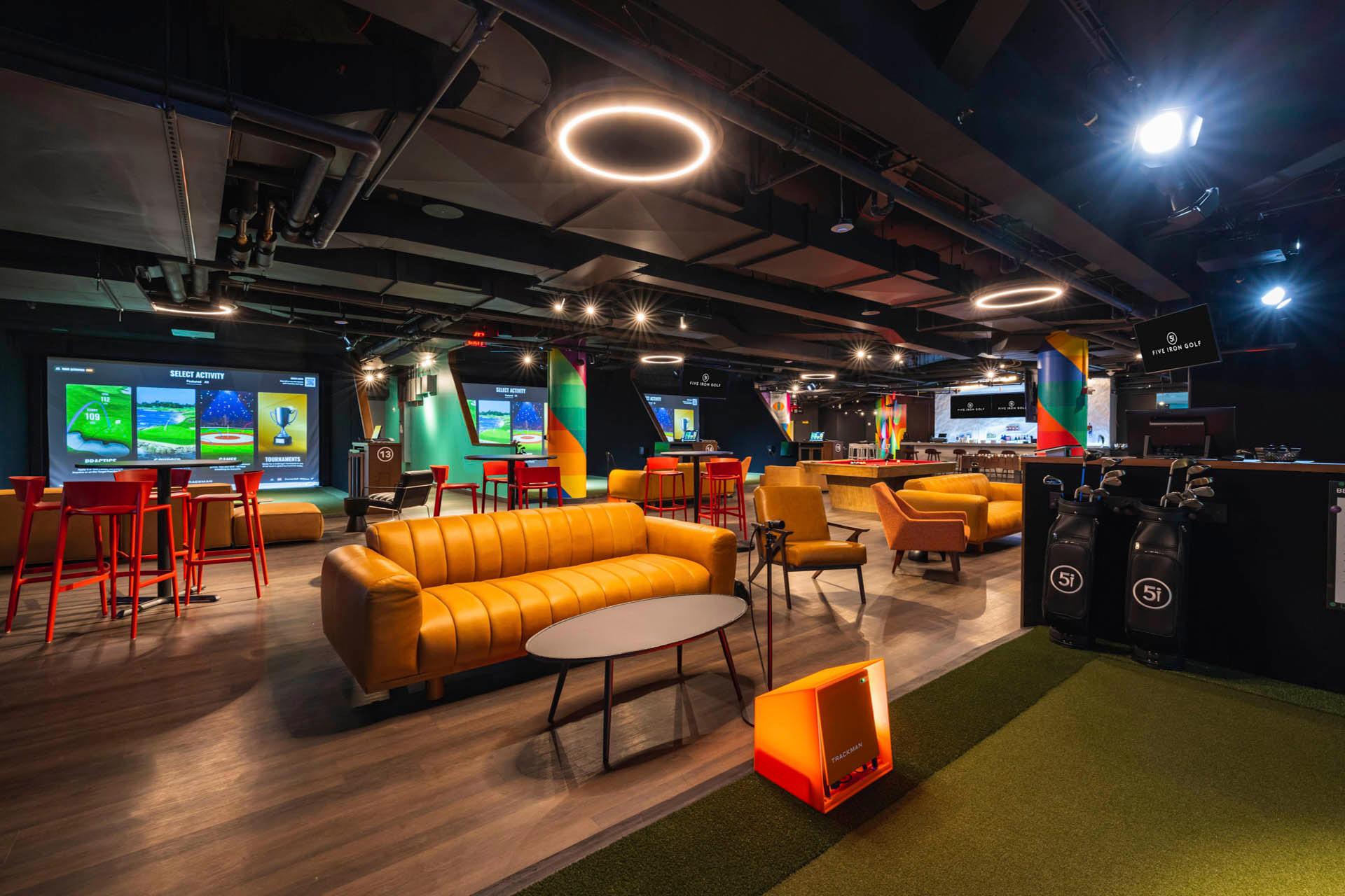 Modern indoor golf lounge with yellow sofas, tall red tables and stools, multiple golf simulators, golf bags, and colorful lighting, creating a relaxed and vibrant atmosphere.