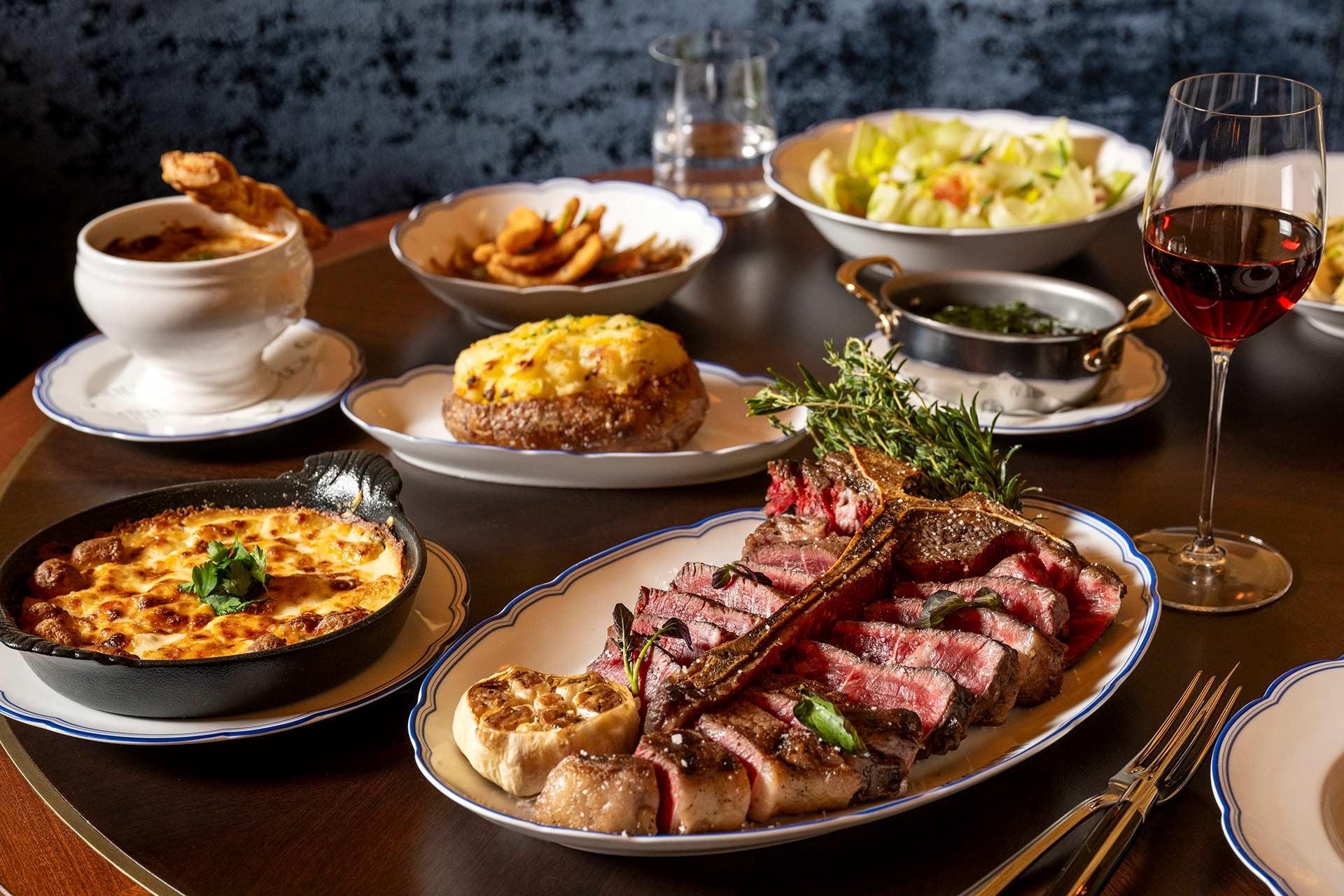 A table set with a sliced steak, baked potato, creamy gratin, French onion soup, salad, sautéed greens, and a glass of red wine. The dishes are attractively arranged on white plates.