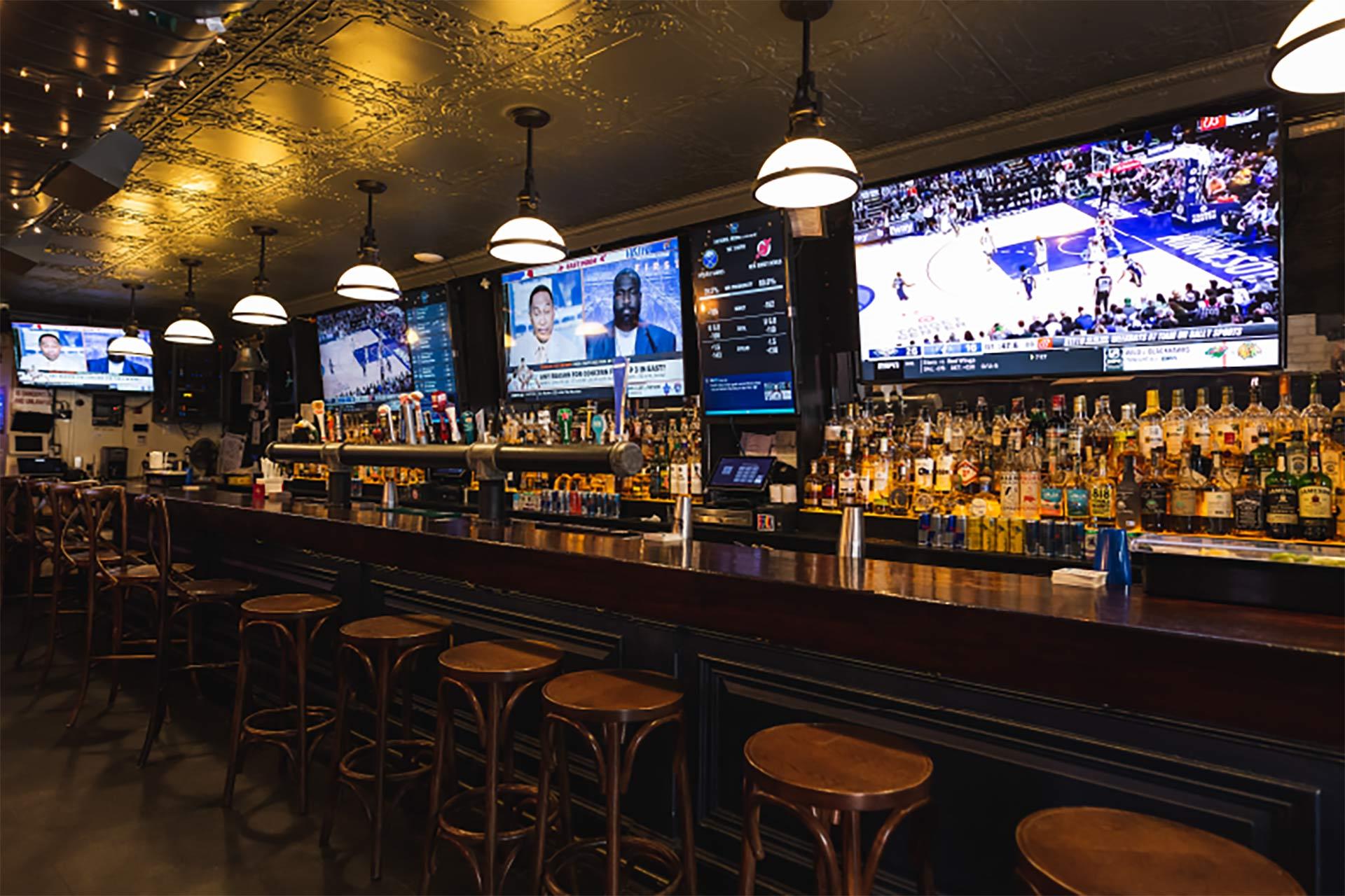 A sports bar with a long wooden counter, rows of empty barstools, shelves of liquor bottles, and several large TVs above the bar displaying news and sports games. Warm pendant lights hang from the ceiling.