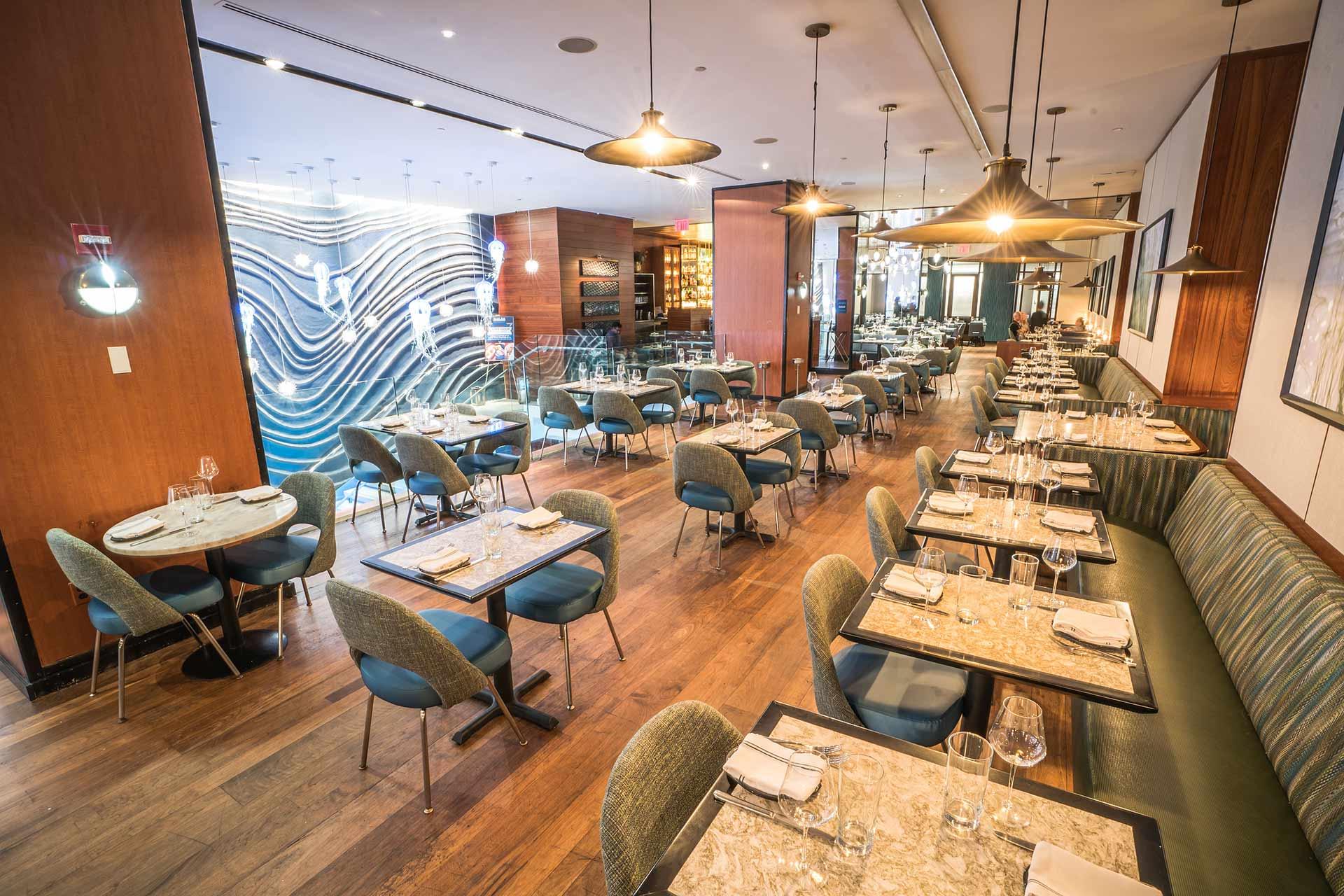 Modern restaurant interior with wooden floors, marble tables, green upholstered chairs, and banquette seating. Stylish lighting hangs from the ceiling, and a wavy blue accent wall decorates the back of the room. Tables are set for dining.