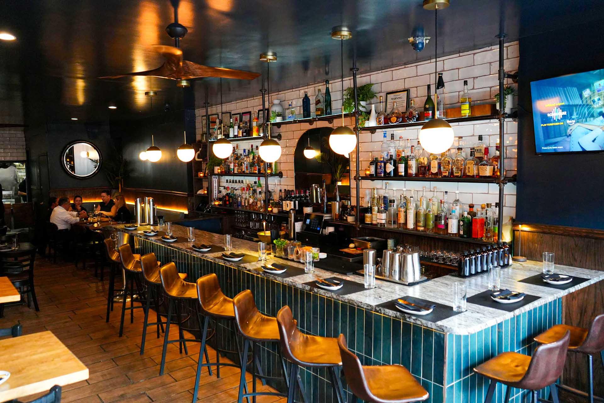 A modern bar with a marble countertop, brown barstools, and shelves of liquor bottles. Warm lighting and a TV on the wall create a cozy atmosphere; a few people are seated in the background.