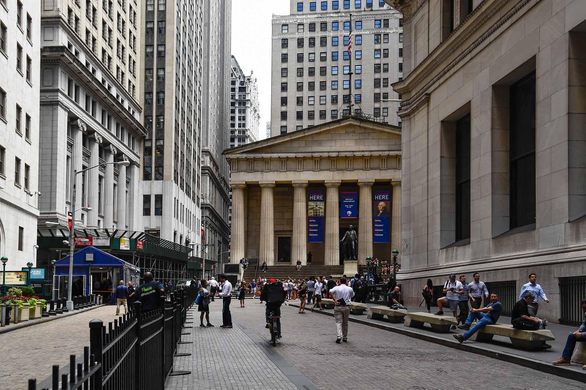 People walk and sit along Wall Street in New York City, with the Federal Hall building and its columns and George Washington statue visible in the background. Tall office buildings line both sides of the street.