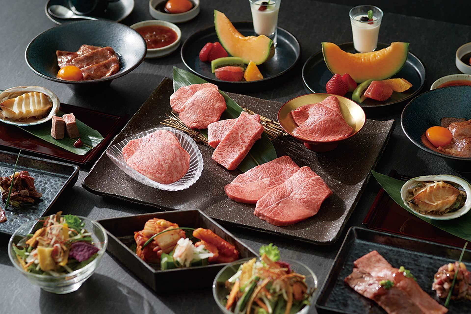 A Japanese meal spread with various dishes, including raw beef cuts, salads, kimchi, fruits, dipping sauces, and desserts, all arranged neatly on a black table.