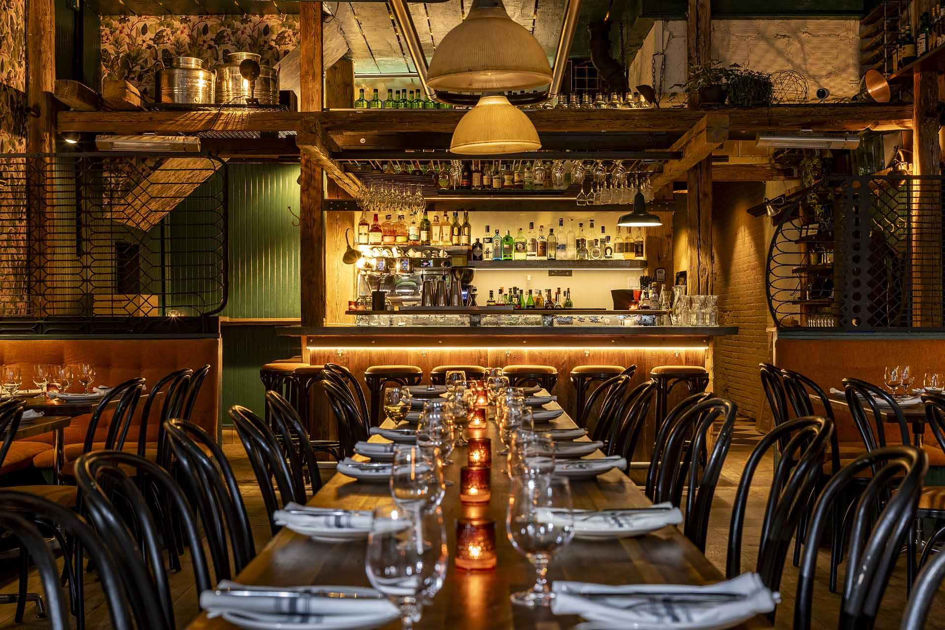 A cozy, dimly lit restaurant interior with long wooden tables set with glasses, plates, and napkins. Candlelight glows along the table. In the background, a well-stocked bar is framed by rustic wooden beams and warm lighting.