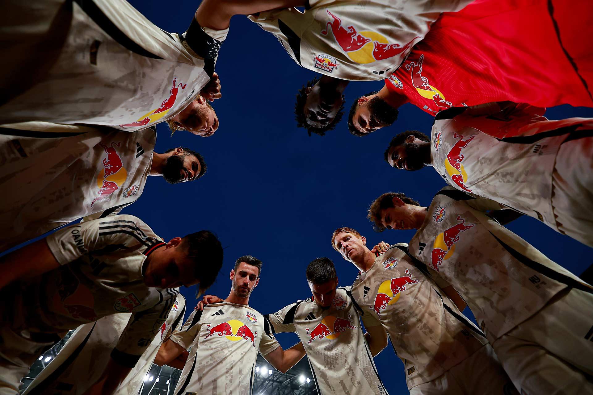 A group of soccer players in light uniforms with a Red Bull logo huddle together in a circle, looking down towards the camera against a dark evening sky.