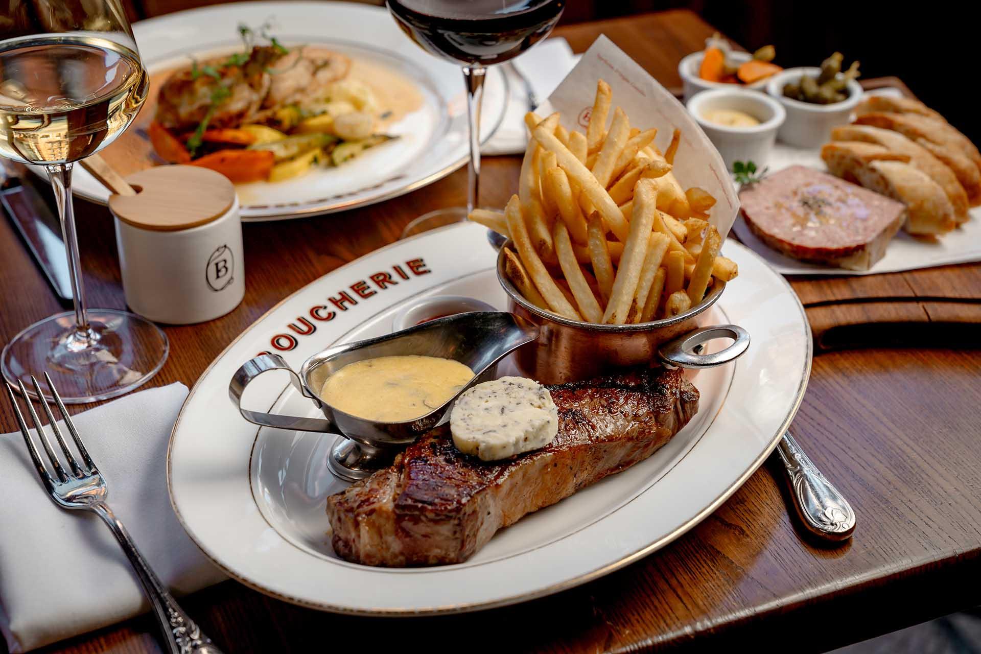 A plate with steak topped with butter, fries in a small pot, sauce, and wine glasses sits on a wooden table. Bread, pâté, and vegetables are in the background.