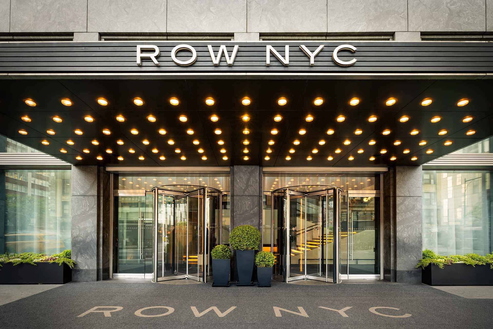 Entrance of Row NYC hotel with rows of lit bulbs on the canopy, revolving glass doors, and large planters with greenery on each side. The hotel's name appears above the entrance and on the doormat.