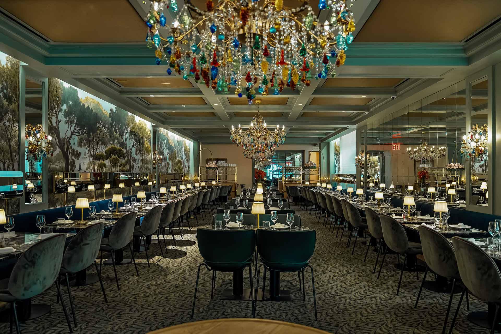 Elegant restaurant interior with long rows of tables set for dining, plush chairs, colorful crystal chandeliers, and large mural-style wall art of trees and landscapes, creating a luxurious and inviting atmosphere.