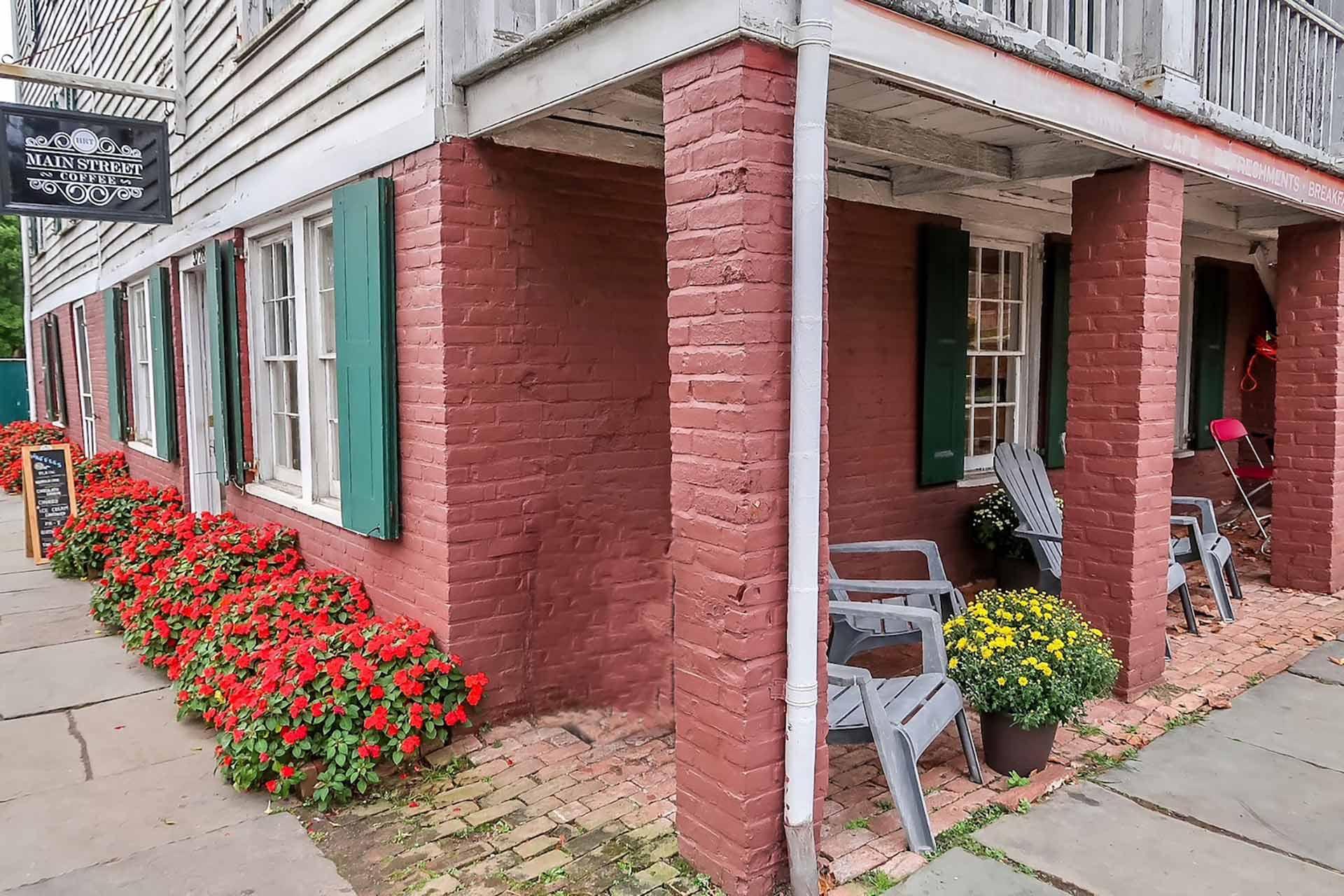 A brick building with green shutters, vibrant red flowers lining the sidewalk, and gray Adirondack chairs with a pot of yellow flowers on a porch under a wooden overhang.