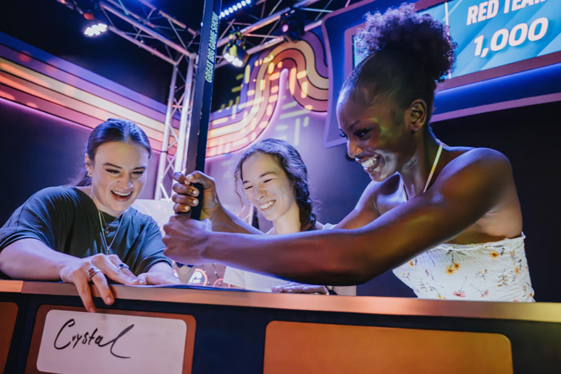 Three women smiling and laughing while participating in an interactive game show activity, standing behind a counter with colorful lights and scoreboards in the background.