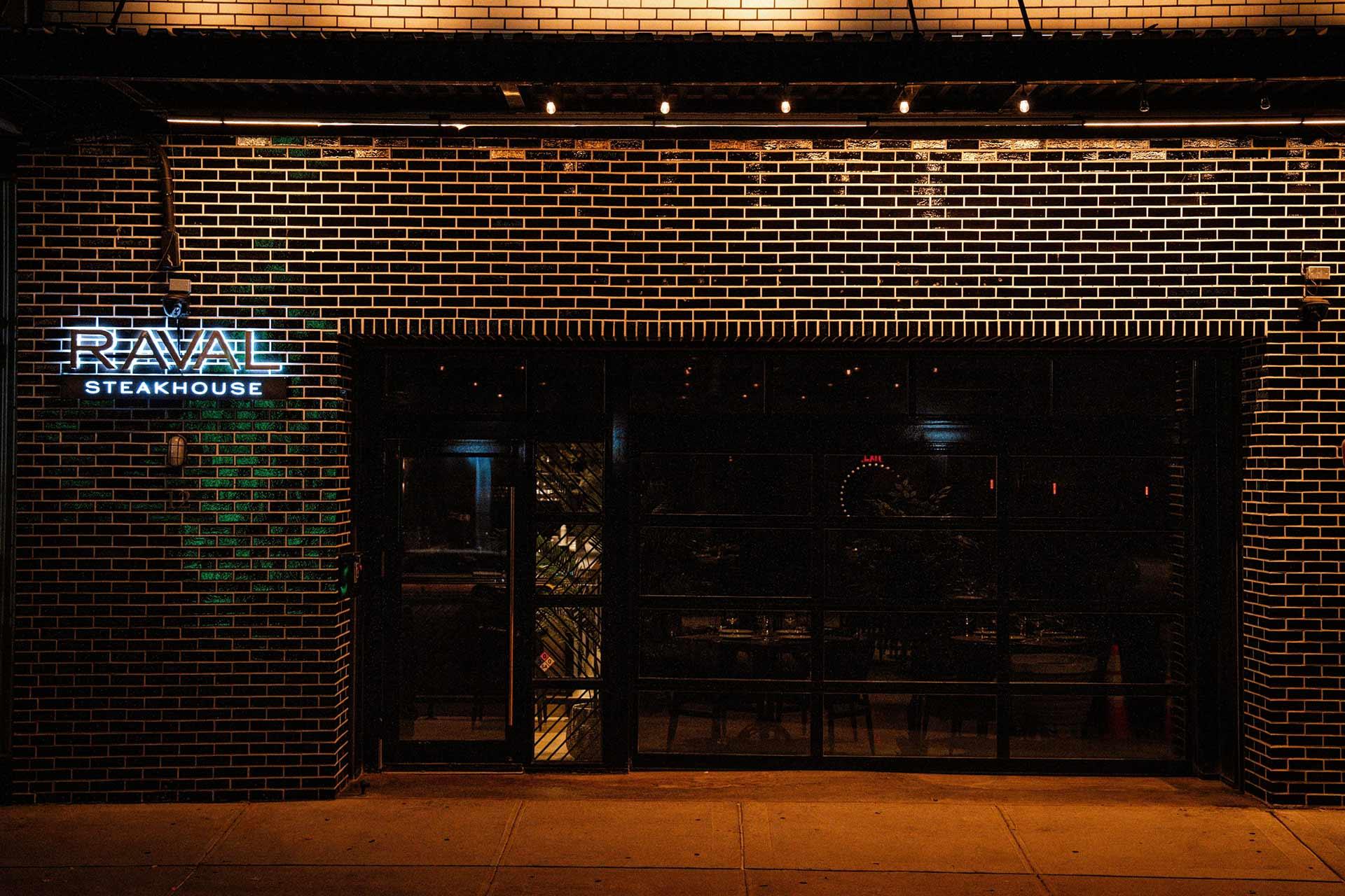 Nighttime view of a brick building with a lit sign reading "RAVAL STEAKHOUSE" on the left, and a large glass door or window revealing a dimly lit interior with tables and chairs inside.