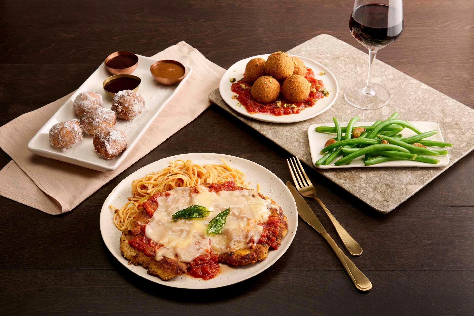 A meal setting featuring a plate of breaded chicken topped with cheese and tomato sauce, served with spaghetti. Surrounding dishes include stuffed balls on tomato sauce, green beans, dessert pastries, and a glass of red wine on a stone platter.