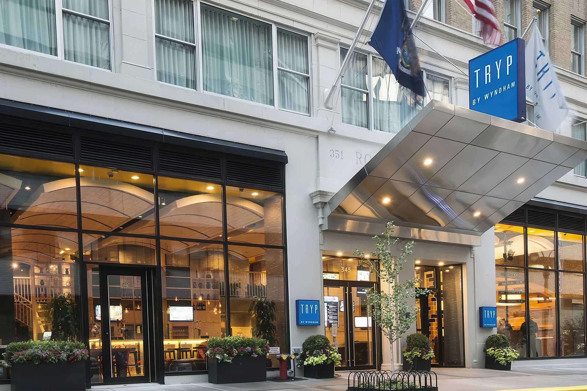 Street view of the entrance to TRYP by Wyndham hotel, featuring large windows showing the modern lobby, a silver canopy, blue hotel signs, and flags above the door.