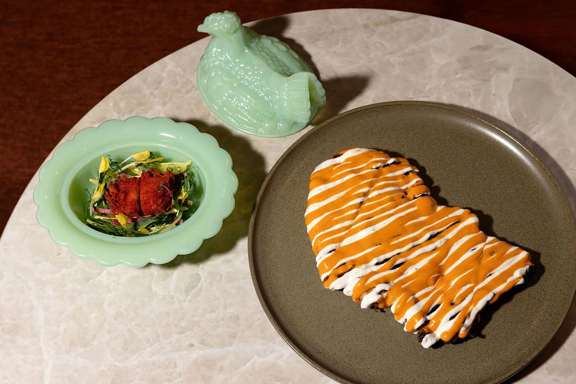 A plate with a large, heart-shaped pastry drizzled with orange and white icing sits next to a green bowl of salad topped with red garnish, and a green ceramic hen container on a marble surface.