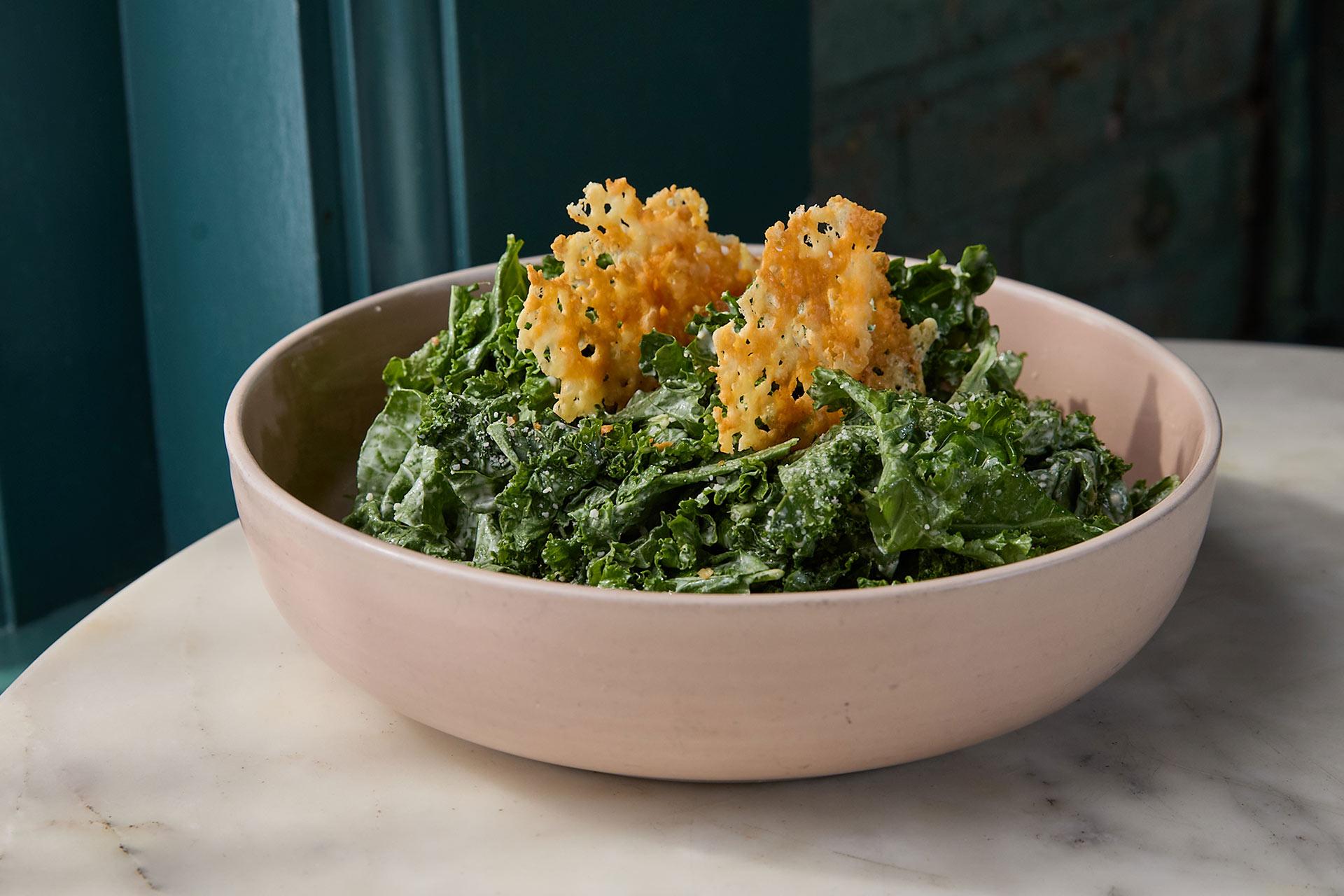A bowl of kale salad topped with two crispy parmesan cheese crisps, served in a light pink bowl on a marble table with a teal wall in the background.