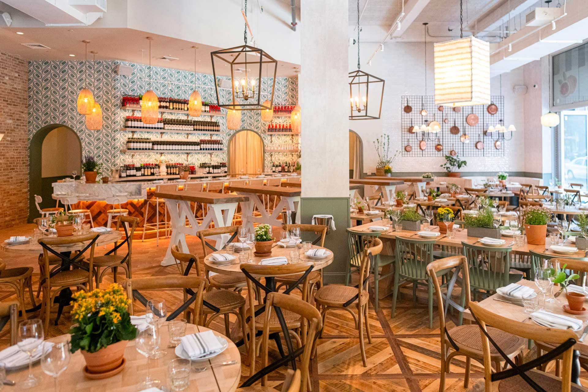 Bright, modern restaurant interior with wooden furniture and patterned tile accents. Tables are set with white napkins and glassware. Potted plants and decorative lights add warmth. Shelves in the background display wine bottles.