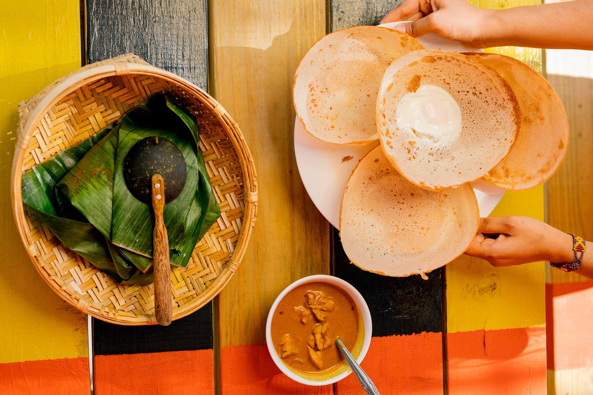 A hand takes a crispy appam with coconut milk from a plate, next to a bowl of curry and a woven basket lined with banana leaves on a colorful wooden table.