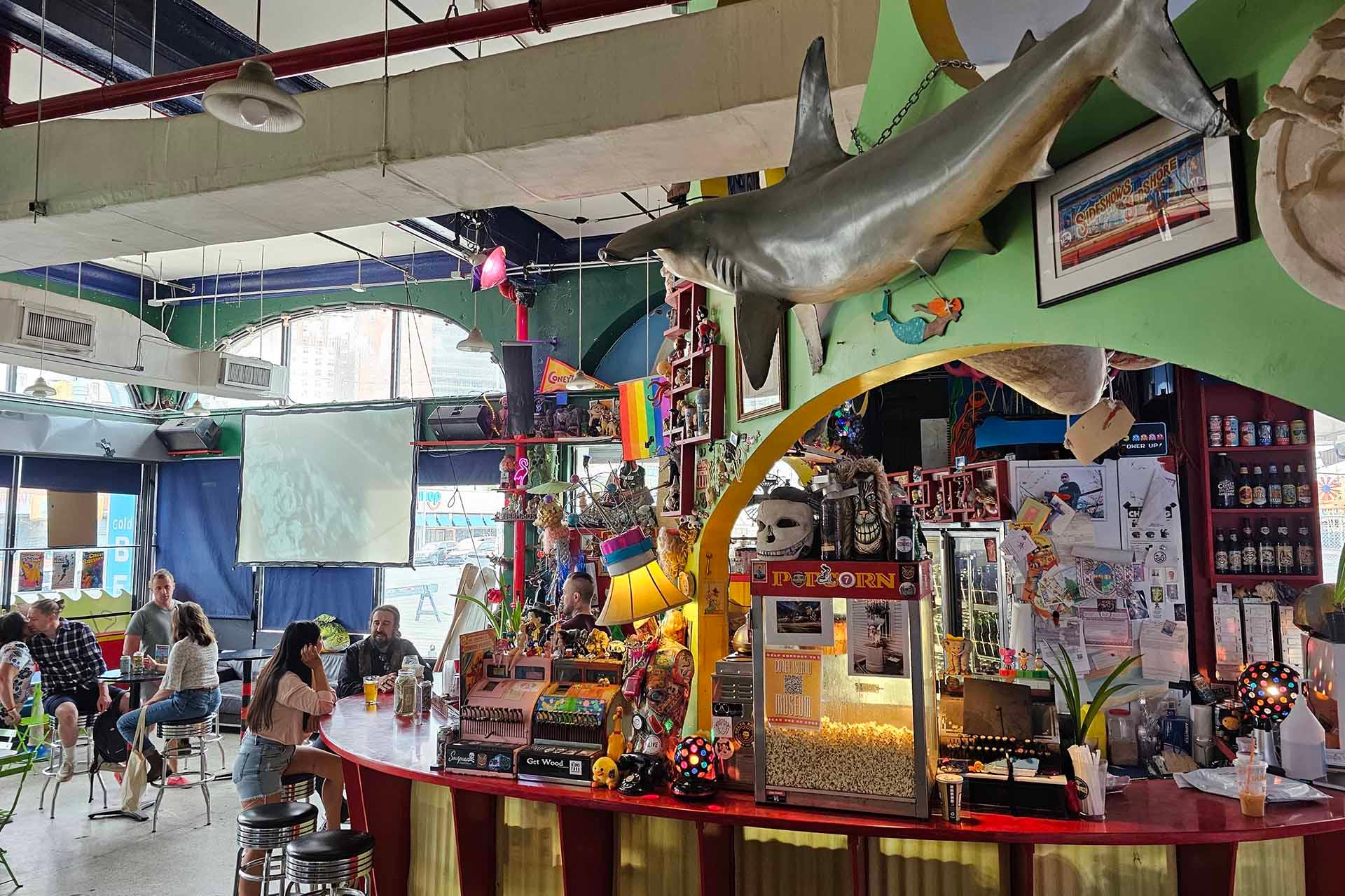 A colorful bar with quirky decor including a large hanging shark, popcorn machine, rainbow flags, and various knick-knacks. Several people sit at tables and the bar, with large windows and a projector screen in the background.