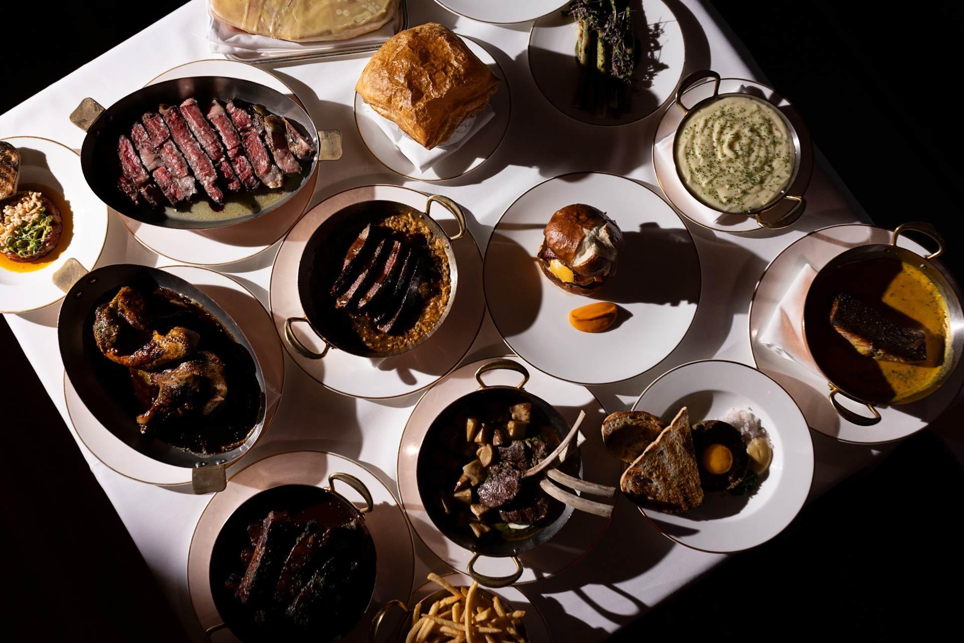 A table set with various plated meats, sauces, mashed potatoes, fries, bread, vegetables, and other sides, all presented in small serving dishes and arranged on a white tablecloth.
