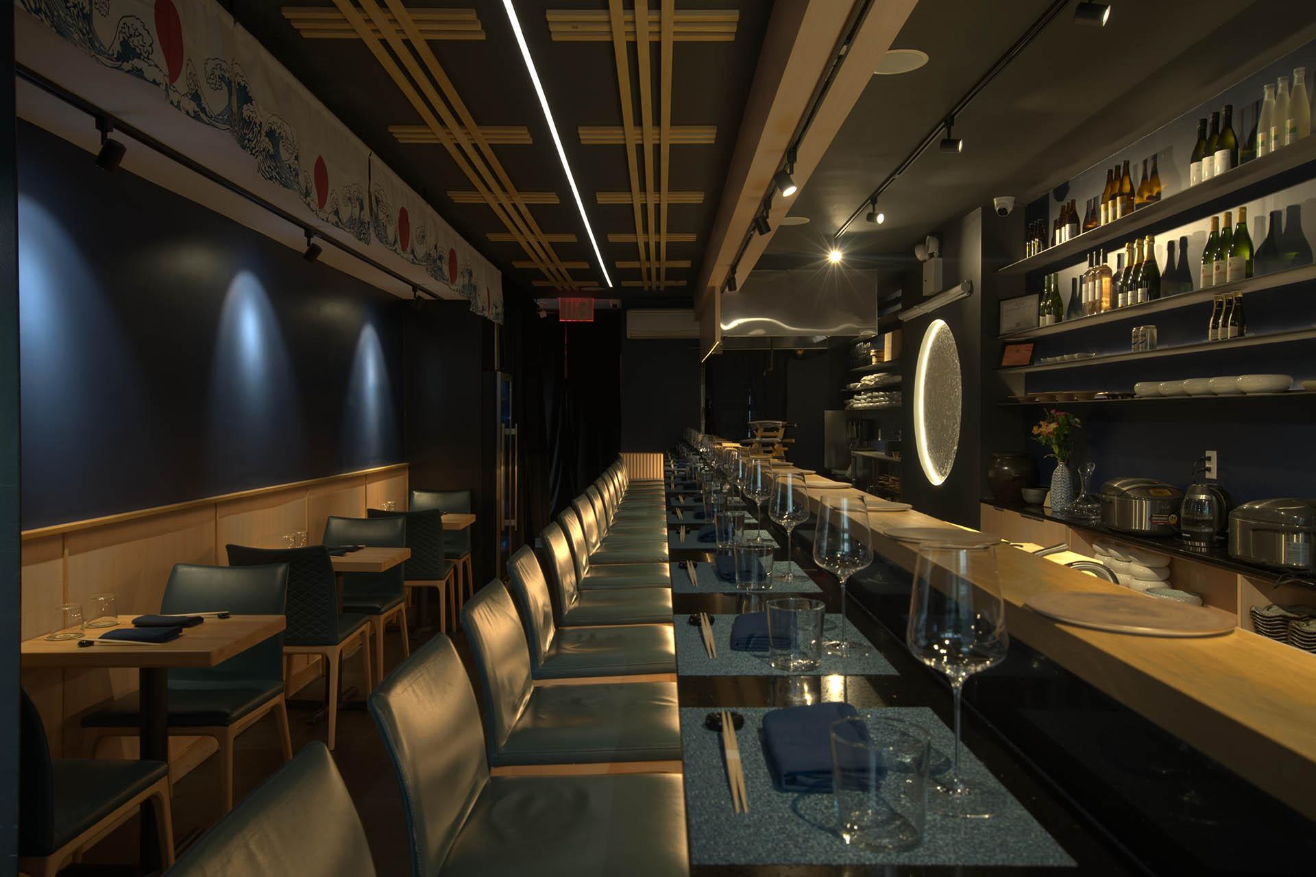 A modern, dimly lit restaurant interior with a long bar, high chairs, neatly set tables, and shelves holding bottles and glassware. The atmosphere is elegant and inviting.