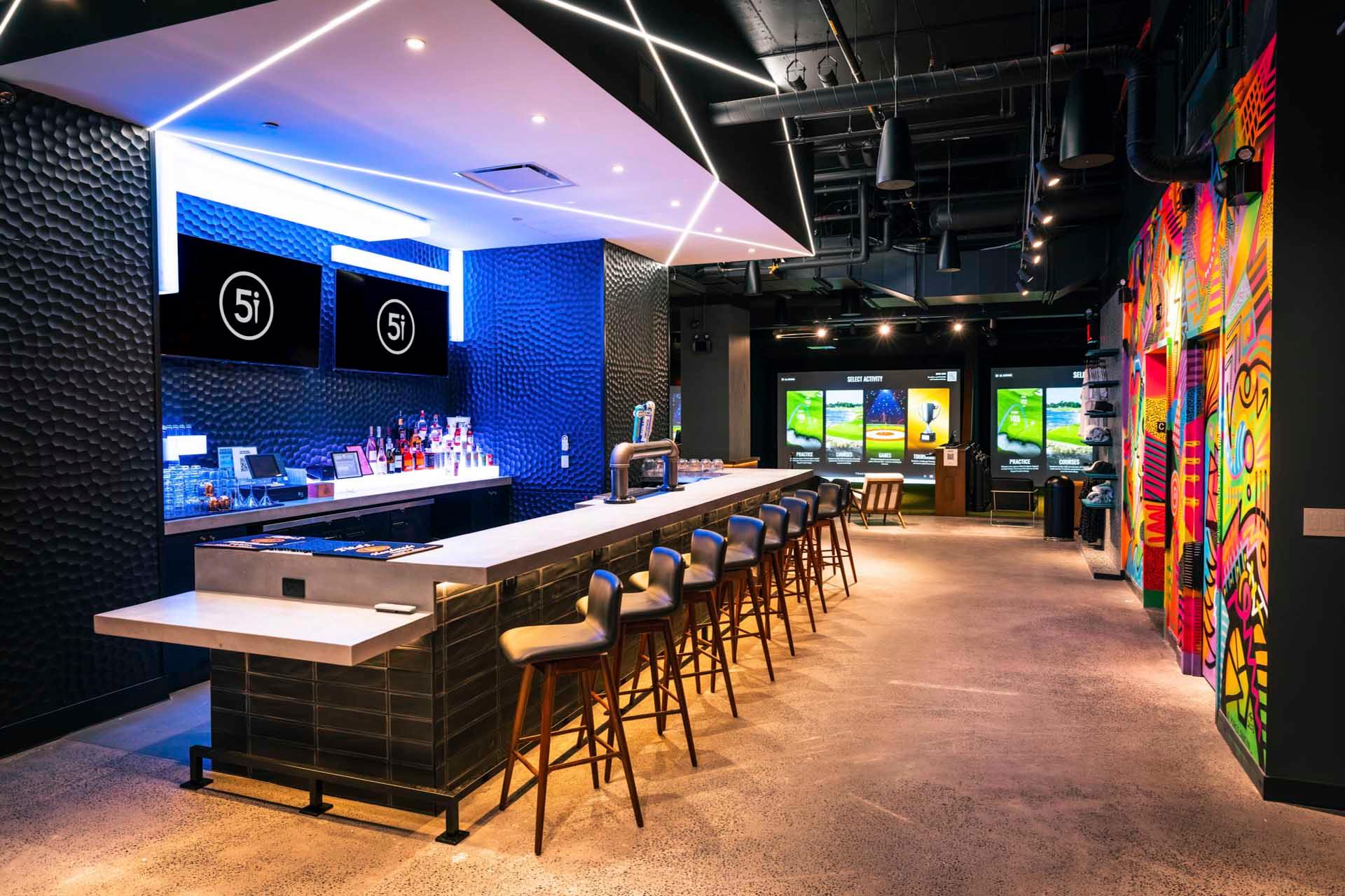 Modern bar with high stools, illuminated blue lighting, and two screens behind the counter. Colorful graffiti art decorates the right wall. In the background, large monitors display golf simulation games. The space is stylish and vibrant.