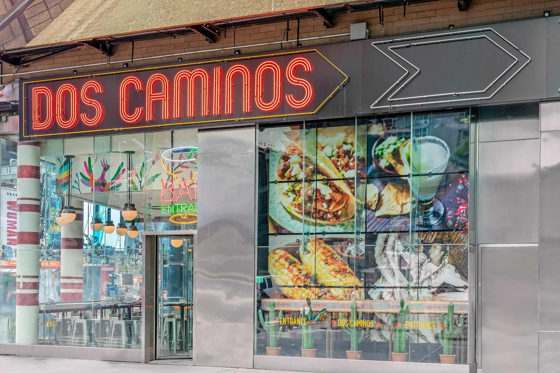 Street view of Dos Caminos restaurant with a neon sign, large arrow, glass windows displaying vibrant food photos, and a modern entrance below additional colorful artwork.