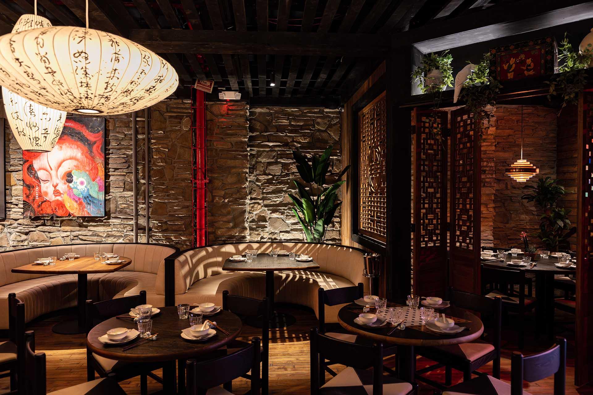 A warmly lit restaurant features stone walls, round wooden tables set for dining, curved beige booths, hanging lanterns with calligraphy, a large colorful painting, and decorative plants, creating an inviting and cozy atmosphere.