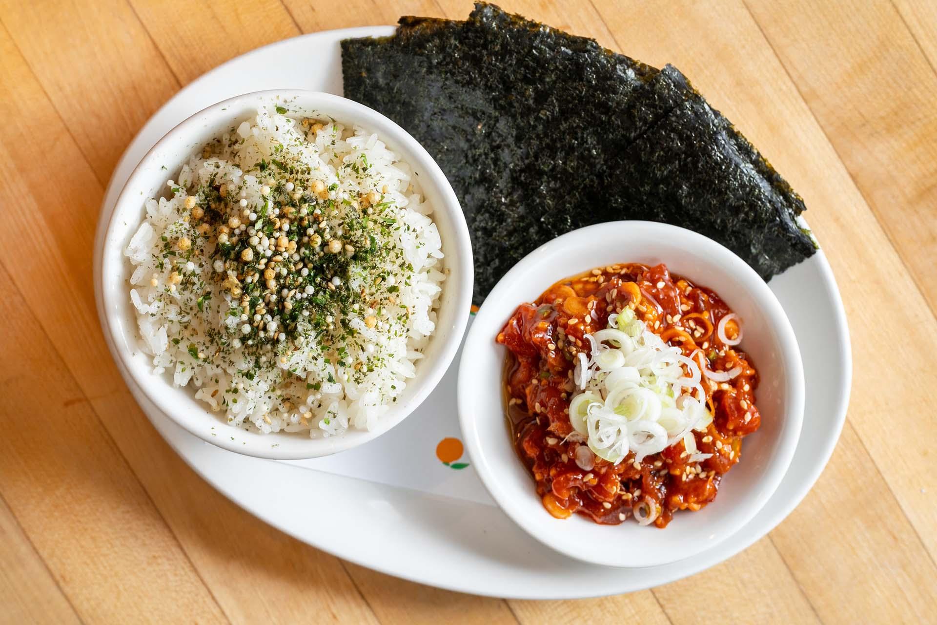 A bowl of white rice topped with seasoning, a bowl of spicy marinated fish garnished with sliced green onions, and several sheets of seaweed on a white oval plate.