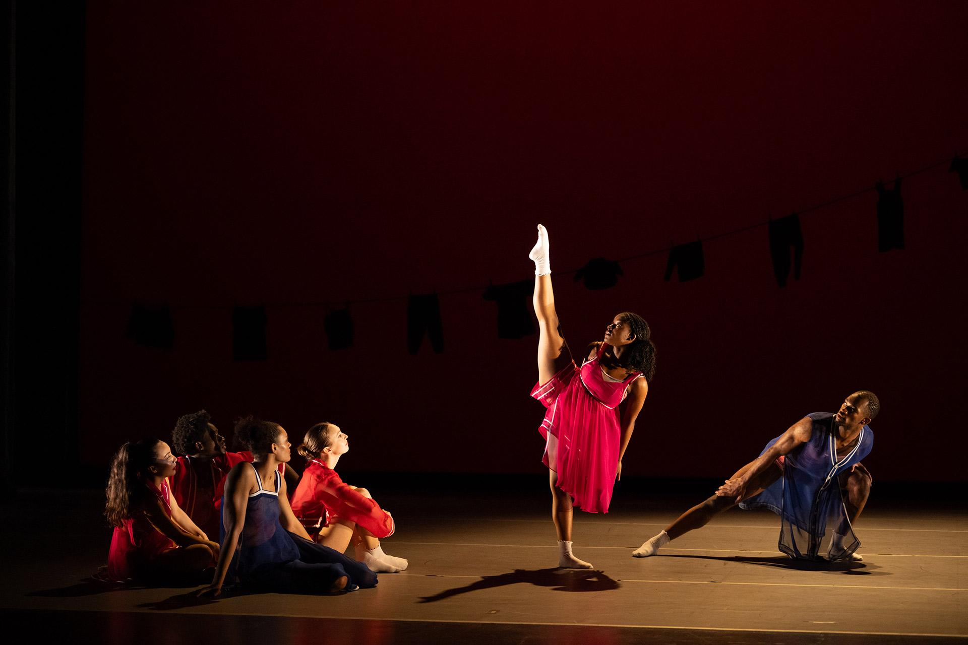 Six dancers perform on stage against a red background. Five are seated or crouching, watching as a woman in a bright pink dress stands with one leg raised high, balancing gracefully under stage lights.