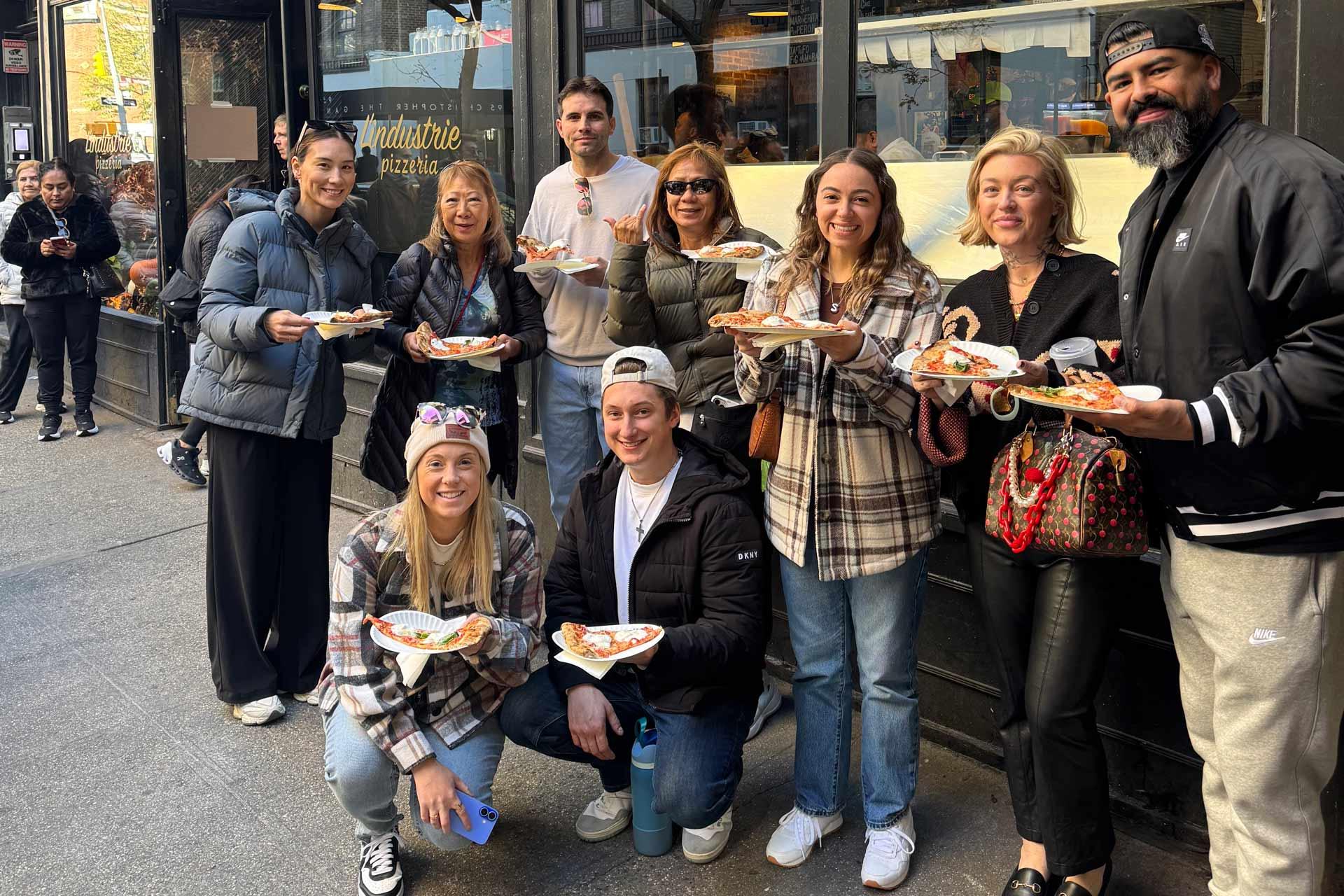 A group of nine people pose outside a pizzeria, smiling and holding plates of pizza. They wear casual fall clothing and stand on a city sidewalk in front of a restaurant window.