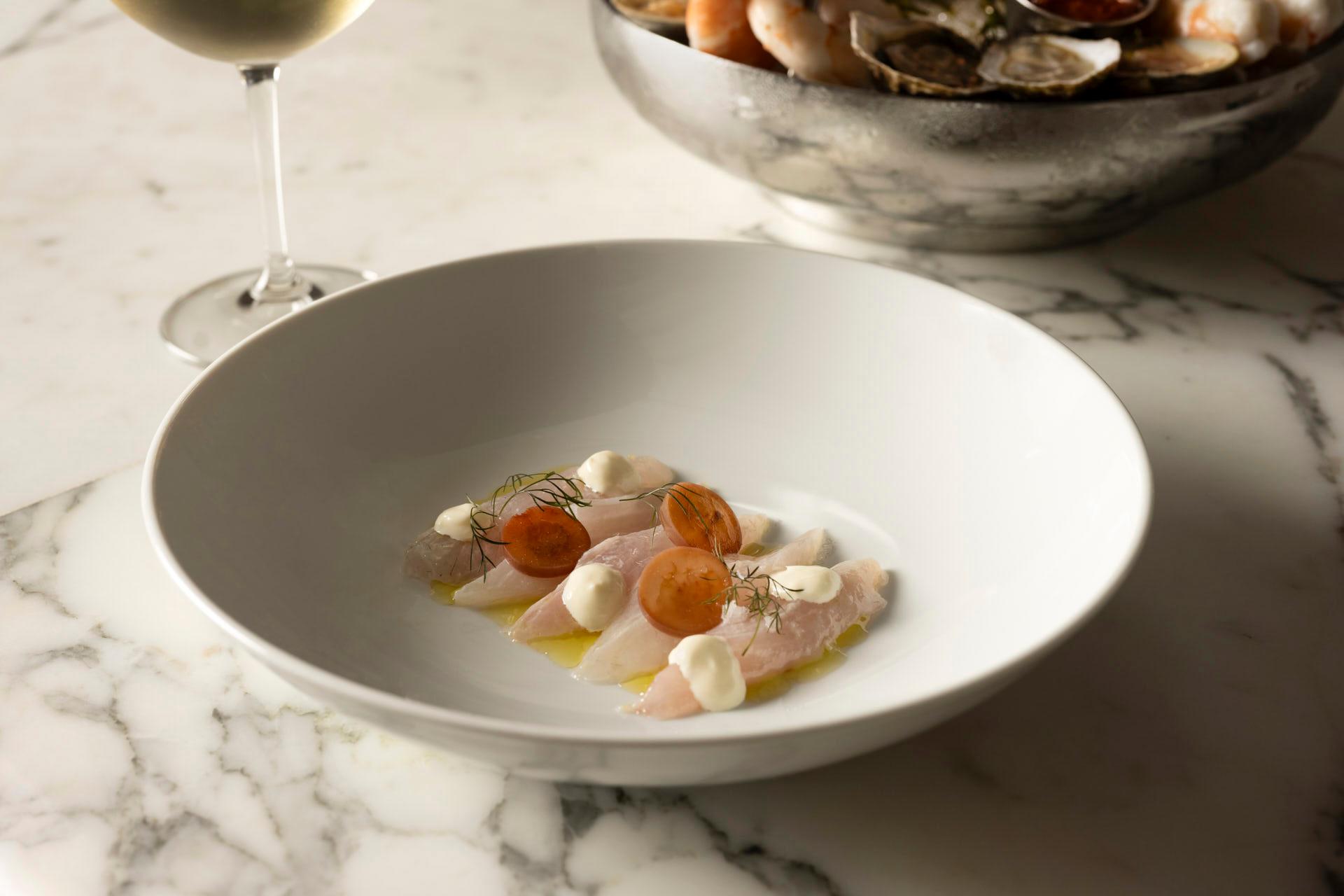 A white plate with thinly sliced fish, topped with small dollops of cream, garnished with herbs and sliced orange vegetables. A glass of white wine and a bowl of seafood are in the background on a marble surface.