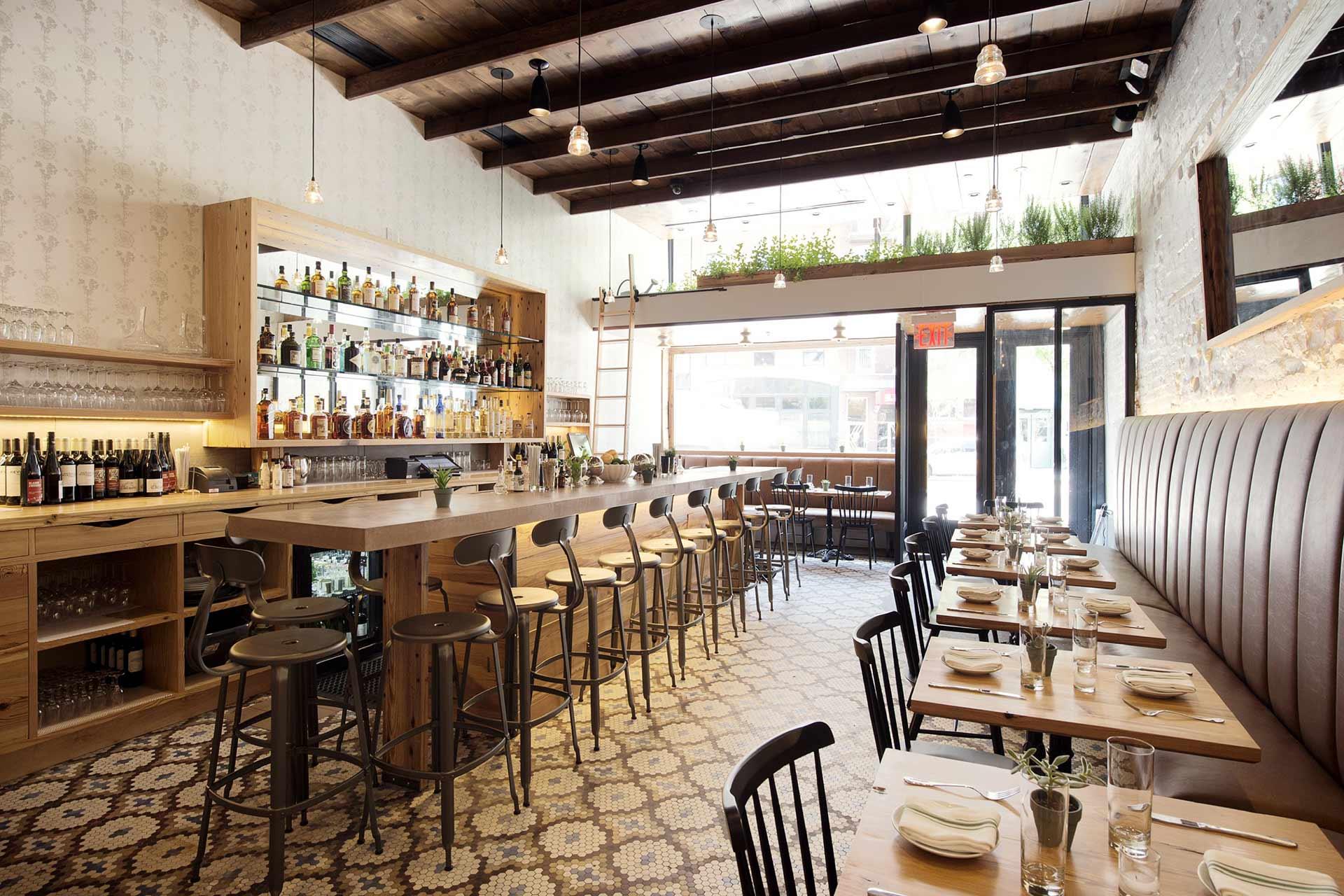 Bright, modern restaurant with wooden tables, metal stools at a long bar, tile floor, and large windows. Neutral tones, bottles on display, and plants on shelves create a welcoming atmosphere.