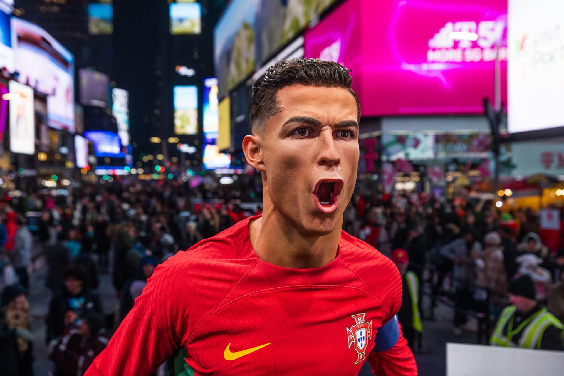 A wax figure of a soccer player in a red Portugal jersey stands with mouth open in Times Square at night, surrounded by crowds and bright, colorful electronic billboards.