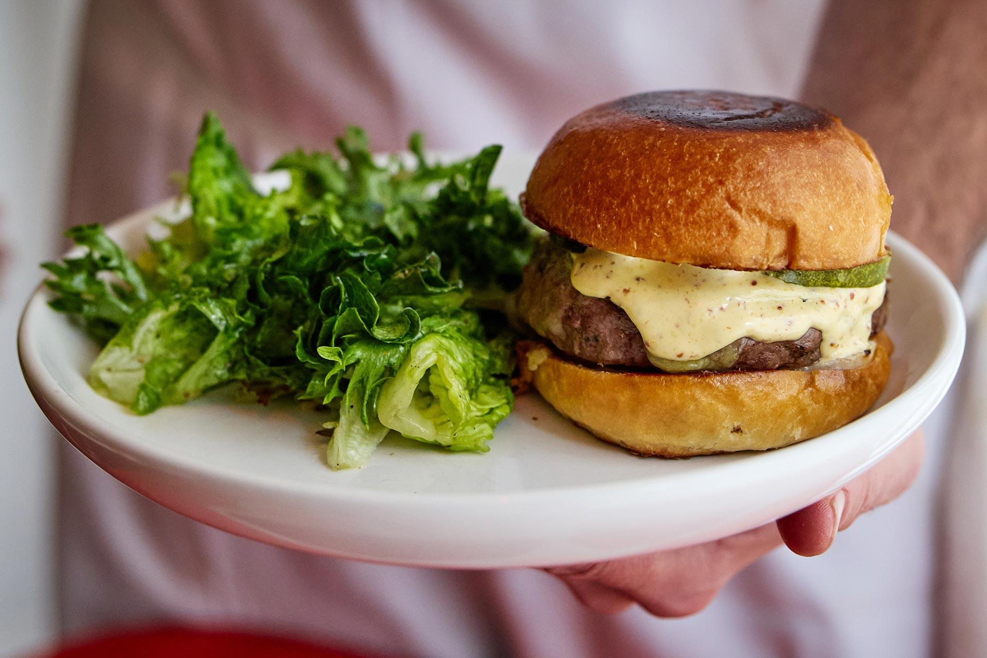 A hand holds a white plate with a cheeseburger topped with pickles and sauce on a toasted bun, served next to a portion of fresh green salad.