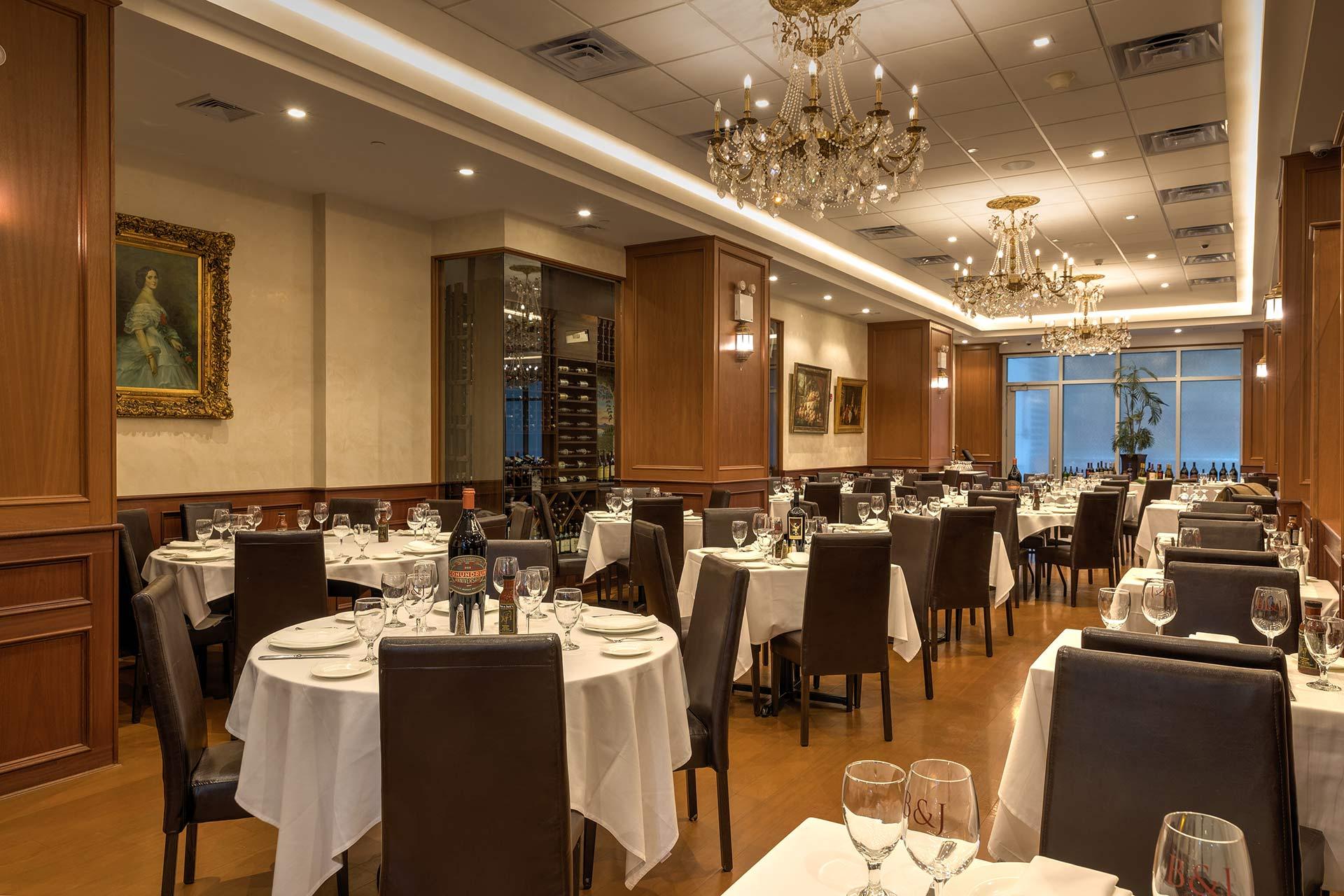 Elegant restaurant dining room with chandeliers, wooden paneling, and tables set with white tablecloths, glassware, and wine bottles. Framed artwork decorates the walls, creating a sophisticated atmosphere.