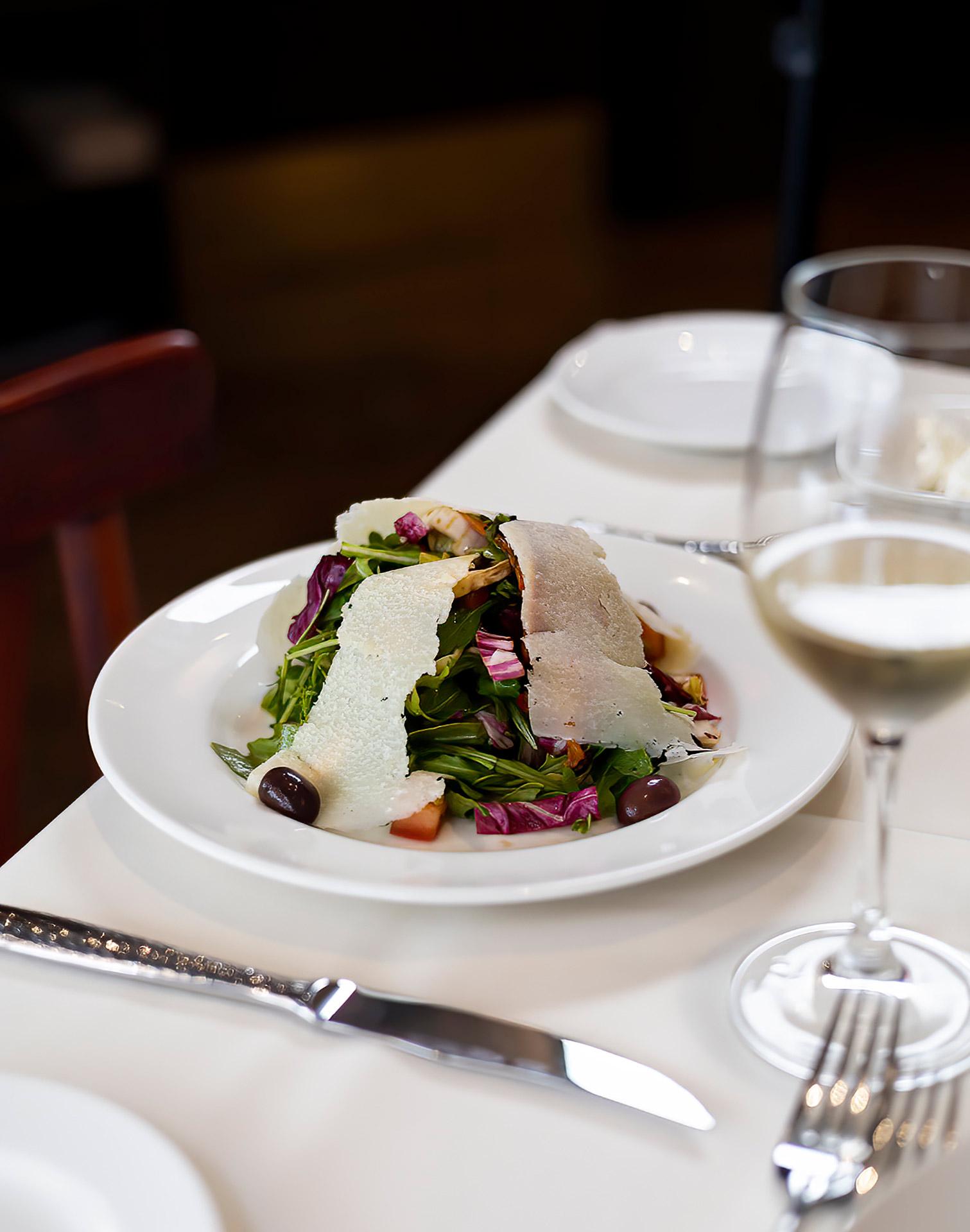 A white plate with mixed greens, olives, tomatoes, and large slices of cheese, set on a white tablecloth beside a wine glass, fork, and knife.