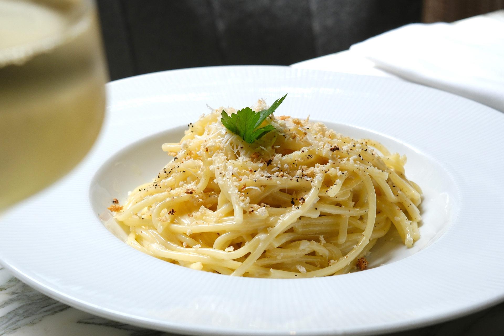 A plate of spaghetti topped with grated cheese, cracked black pepper, and a sprig of parsley sits on a white tablecloth, with a glass blurred in the foreground.